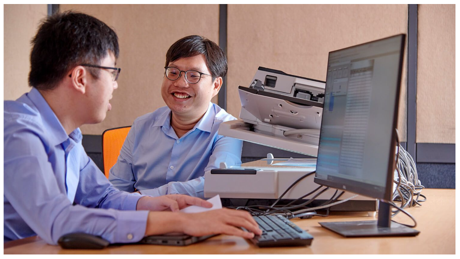 Two men in blue shirts sit at a desk with a computer monitor, keyboard, and document scanner, one smiling.