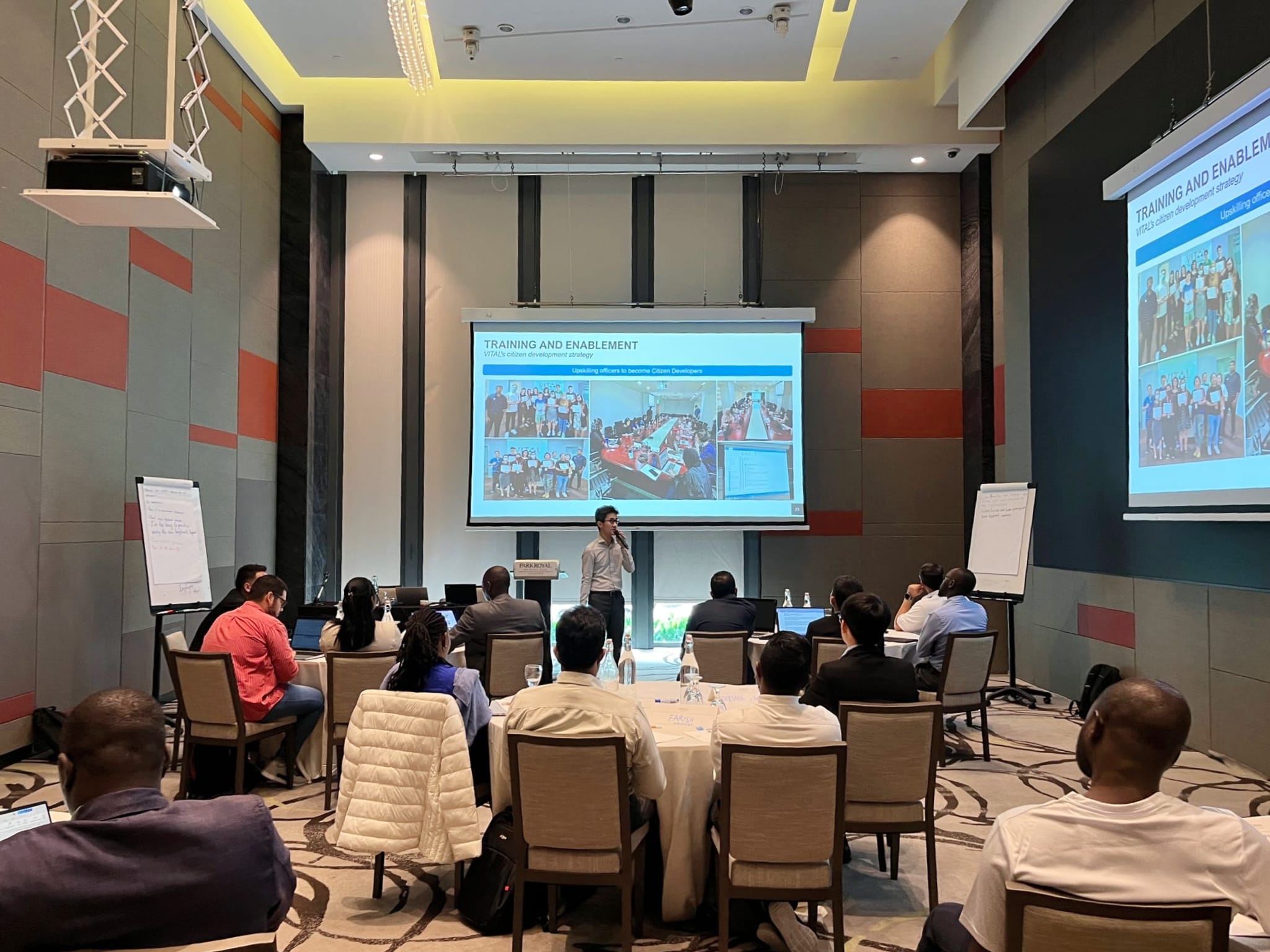 A man presents a "Training and Enablement" slideshow to attendees seated at round tables in a conference room.