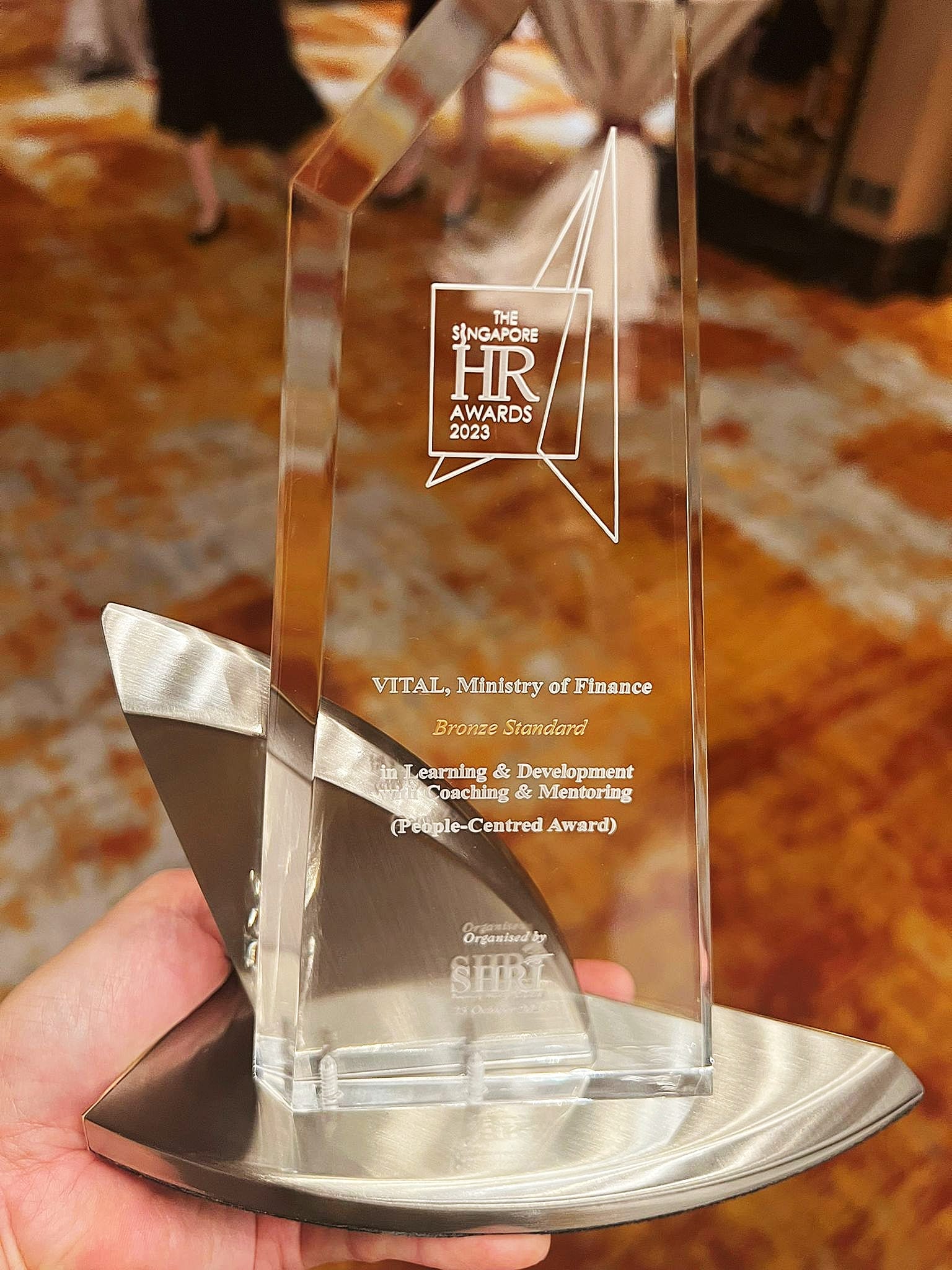 A hand holds a glass trophy for The Singapore HR Awards 2023, Bronze Standard, awarded to VITAL, Ministry of Finance.