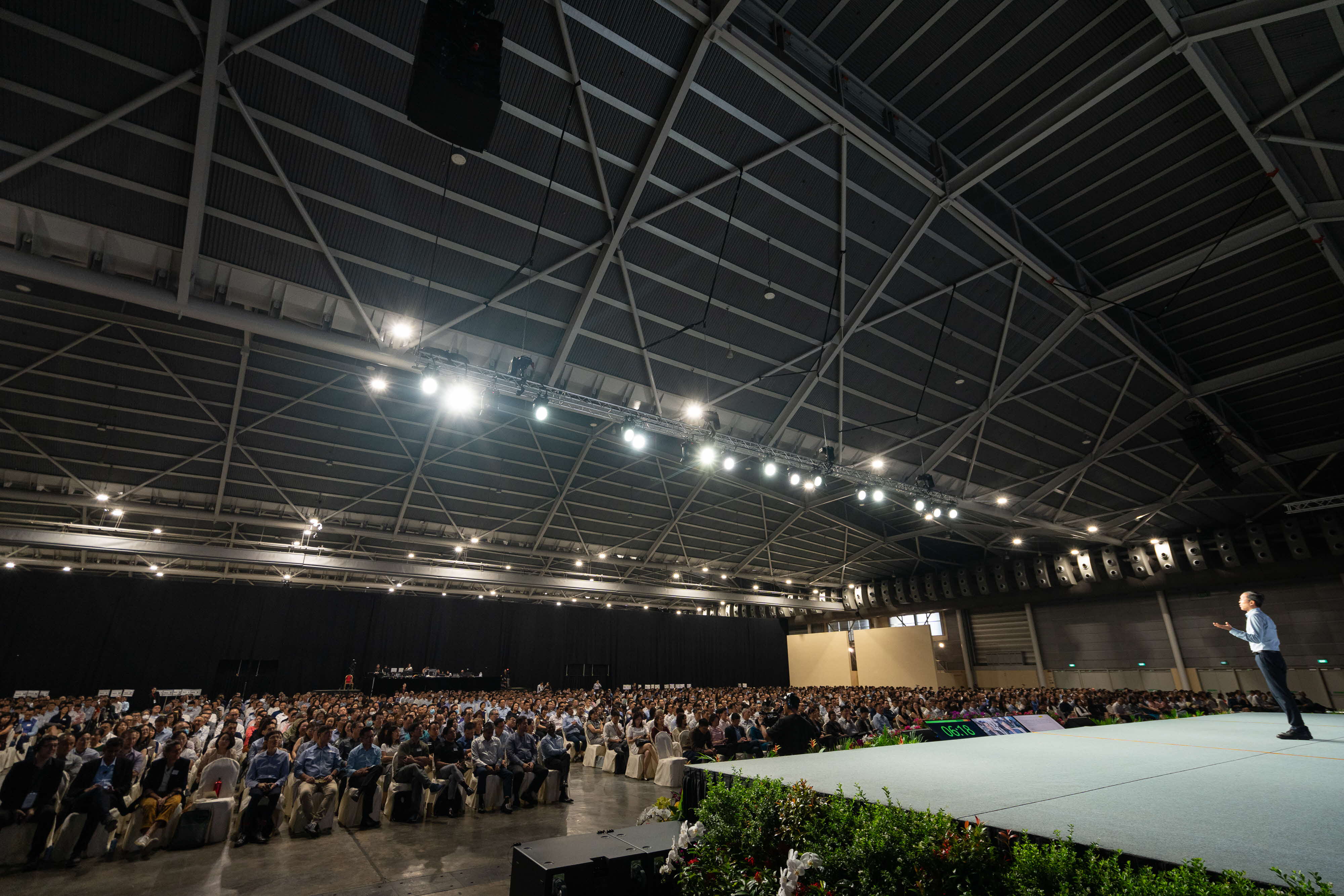 A speaker stands on a lit stage addressing a large seated crowd in a vast indoor hall with steel truss ceiling.