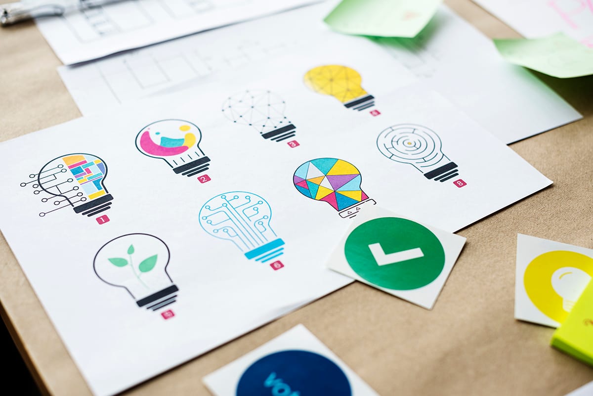 Printed sheets on a desk showing eight numbered colorful lightbulb icon designs, with stickers and sticky notes nearby.
