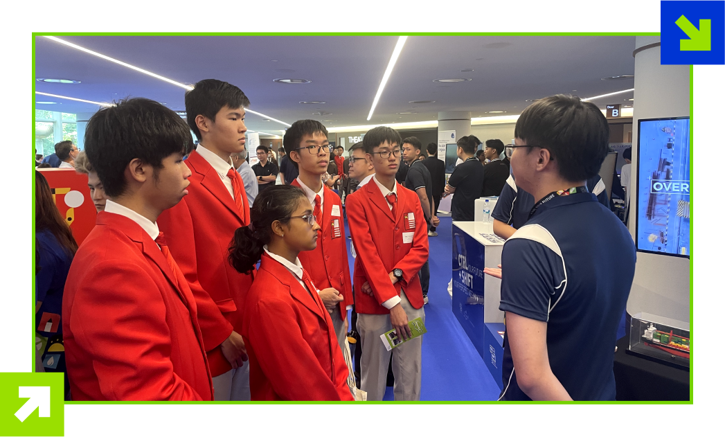 Students in red blazers listen to a presenter at an indoor exhibition booth with screens and displays.