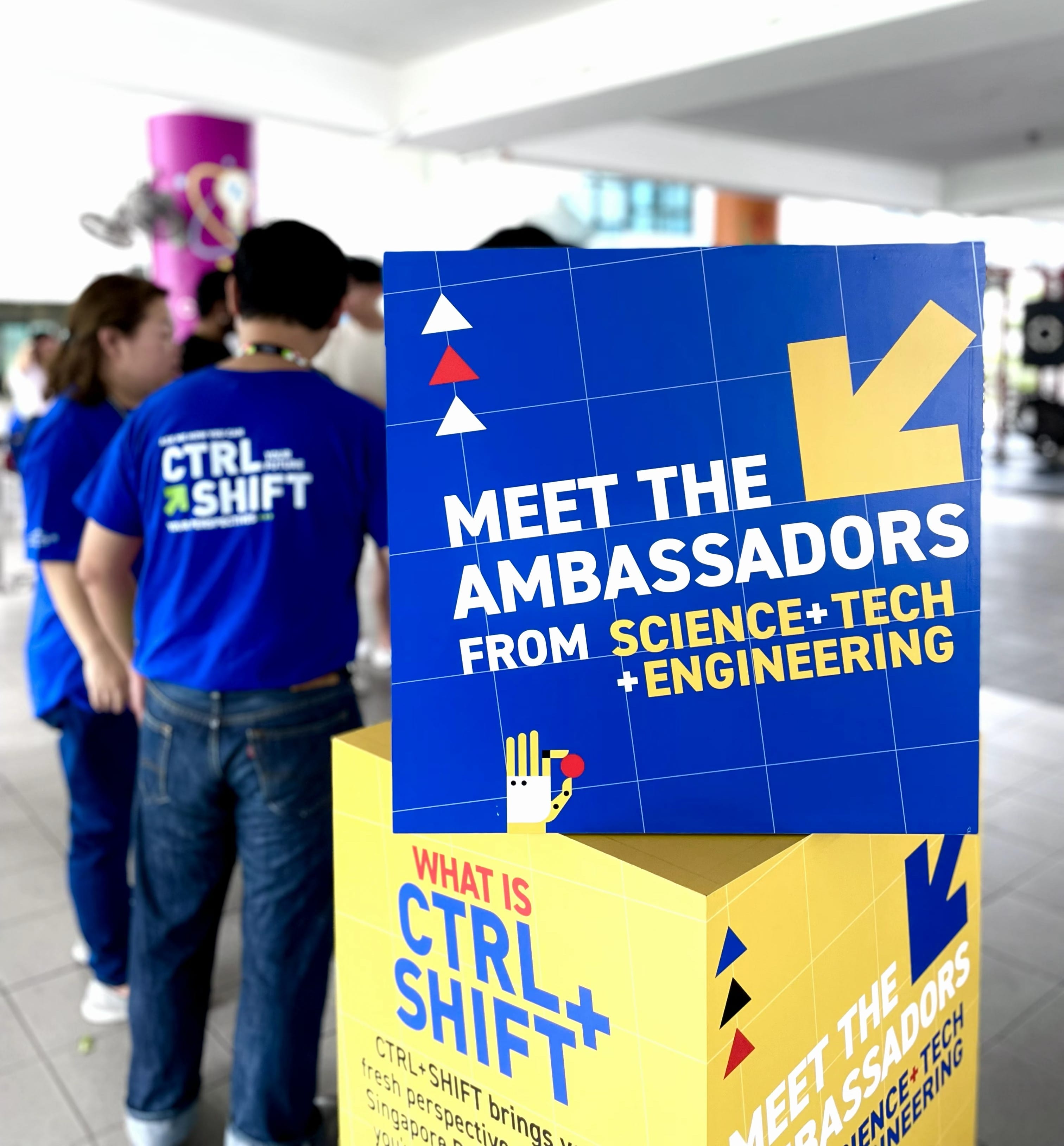Blue sign reads "Meet the Ambassadors from Science+Tech+Engineering" with people in blue shirts standing nearby.