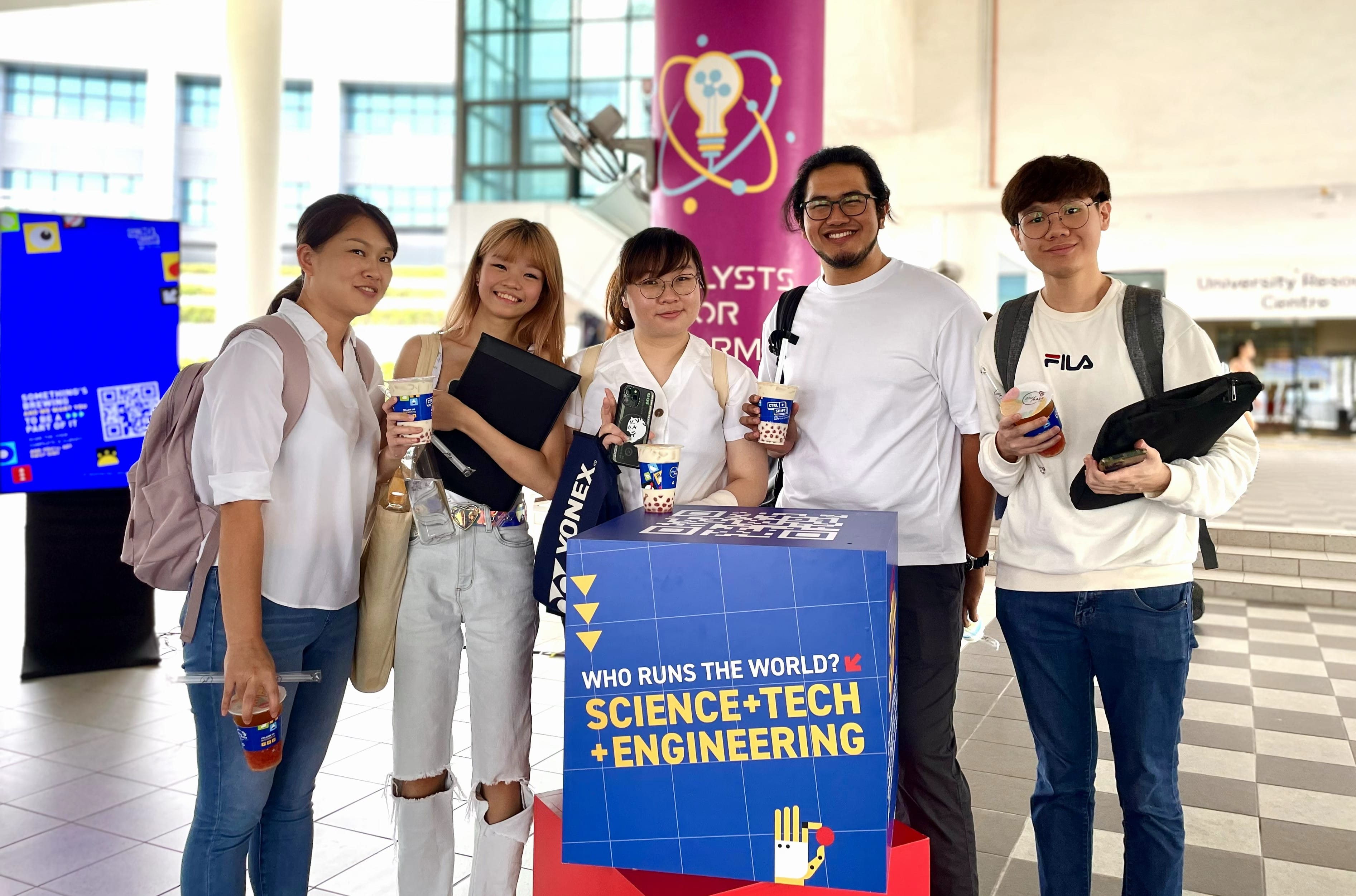 Five young people stand around a blue table reading Science, Tech, and Engineering in front of a pink banner.