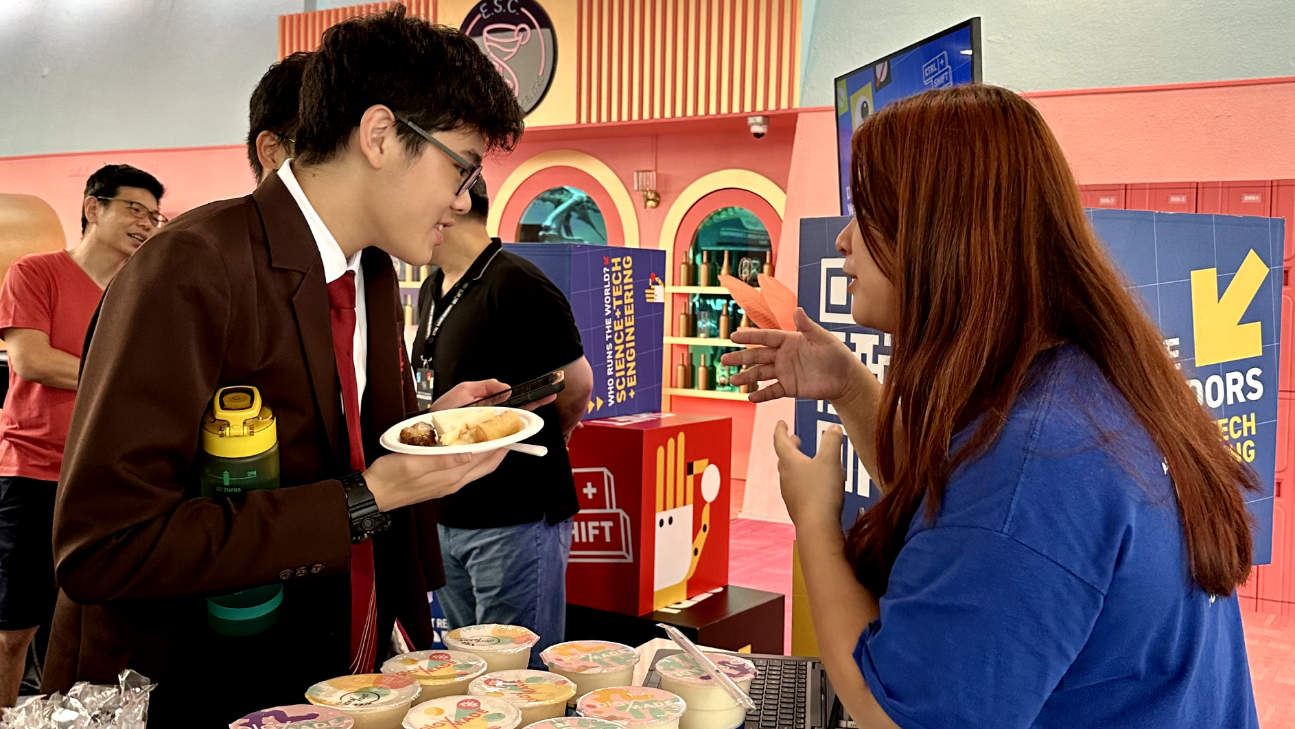 Two people chat at an indoor event booth, one holding a plate of food, with colorful displays in the background.