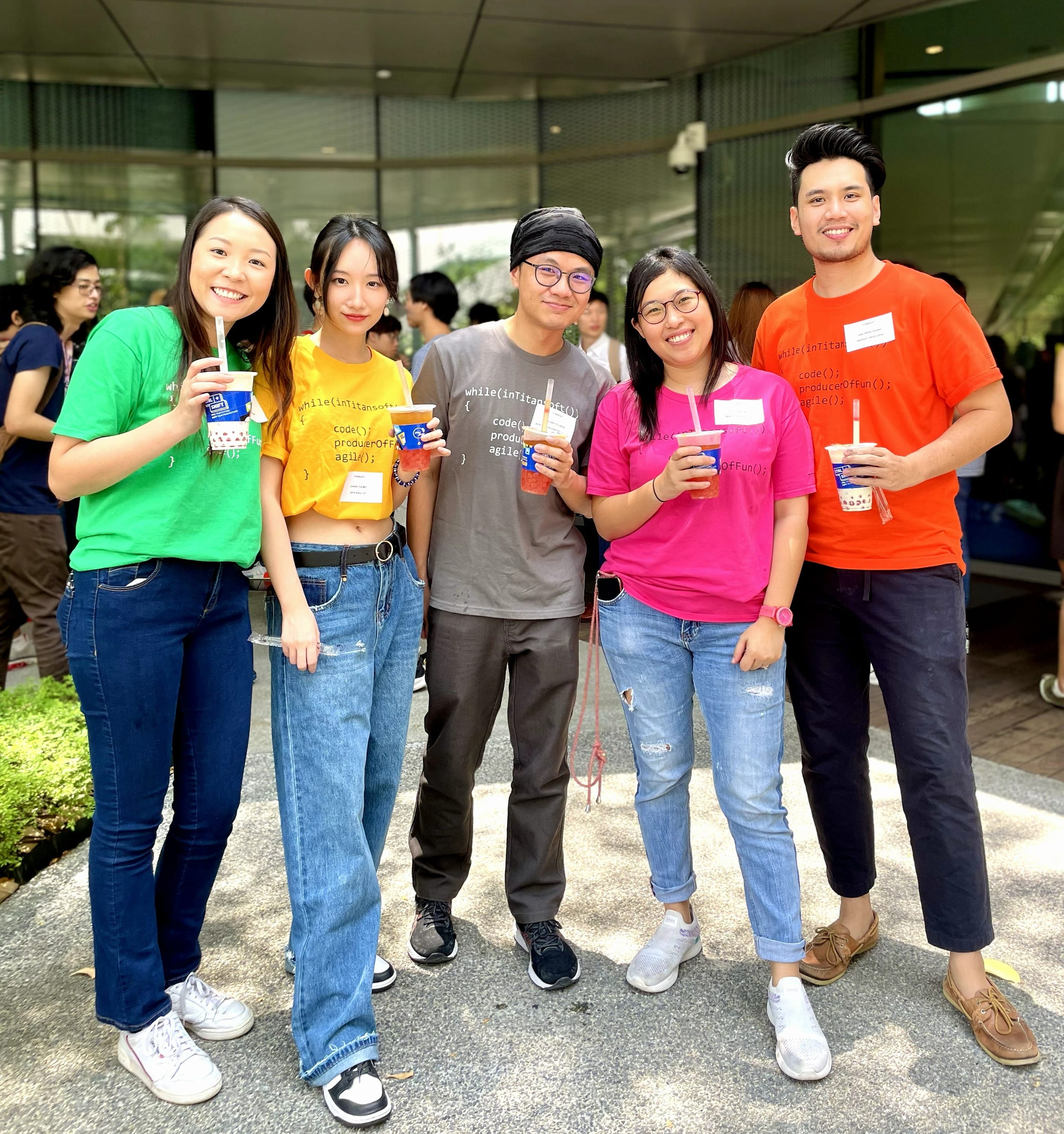 Five young adults wearing colorful shirts smile together outdoors, each holding a blue and white cup.
