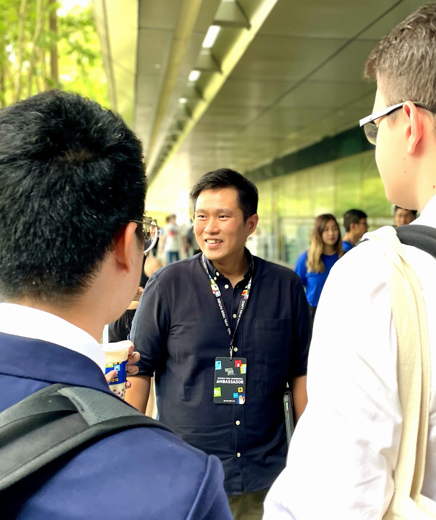 A smiling man wearing a dark shirt and lanyard stands talking with people in an outdoor public area.