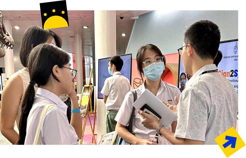 Students in school uniforms converse at an indoor exhibition, one wearing a face mask and holding papers.