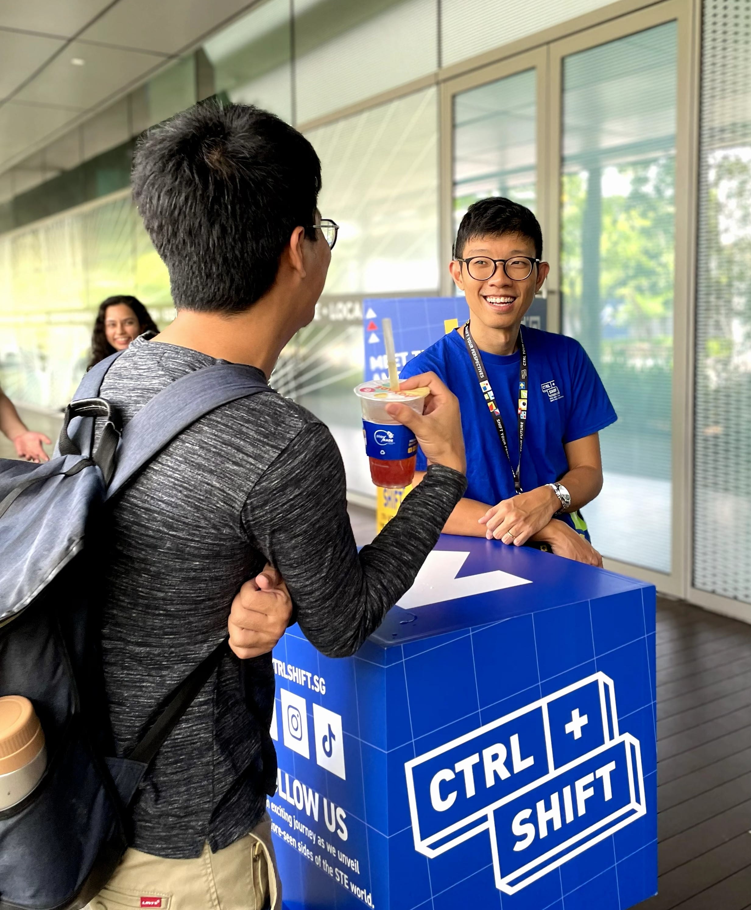 Smiling young man in blue shirt and lanyard hands a drink cup to another person at an outdoor booth.