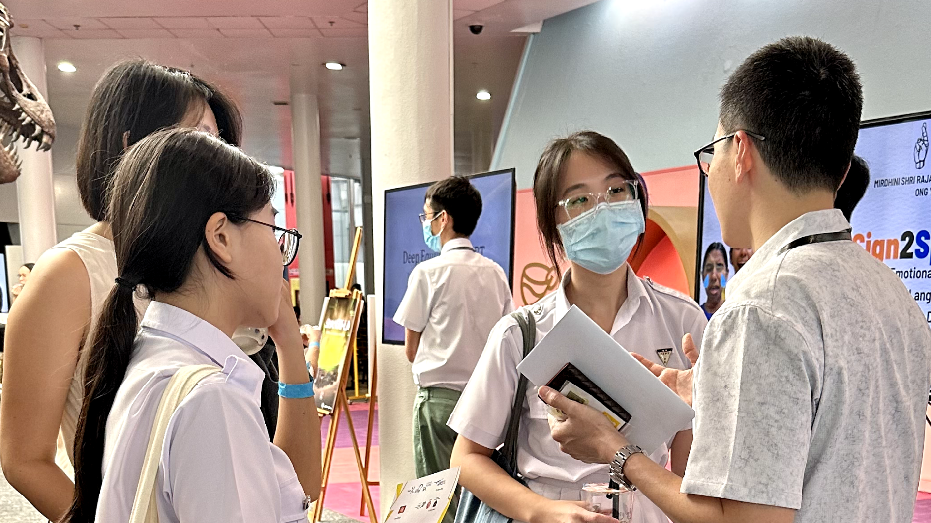 Three students in white uniforms chat at an indoor event, one wearing a face mask and holding folders.