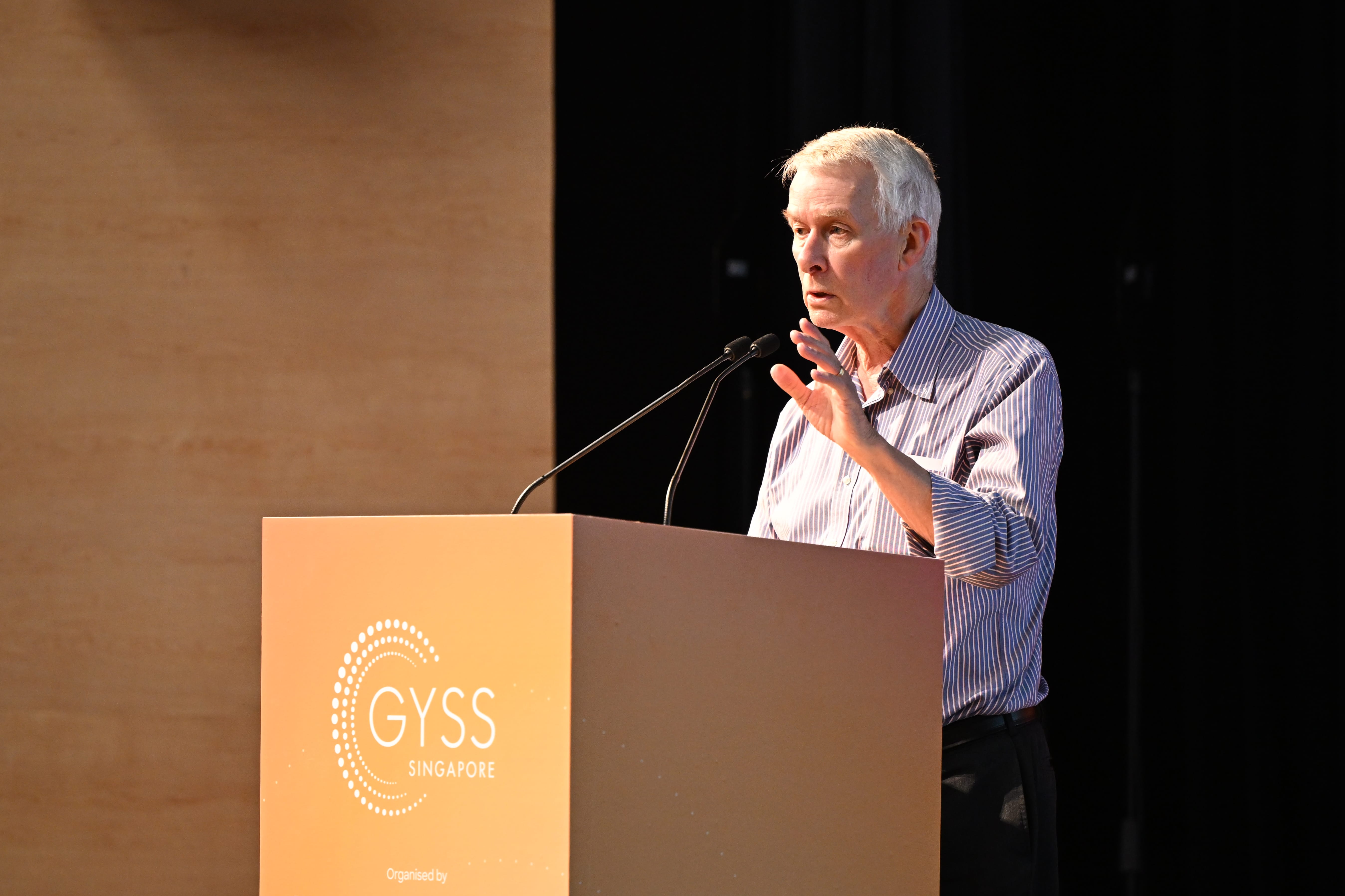An elderly white-haired man speaks at a GYSS Singapore podium with a microphone, gesturing with one hand.
