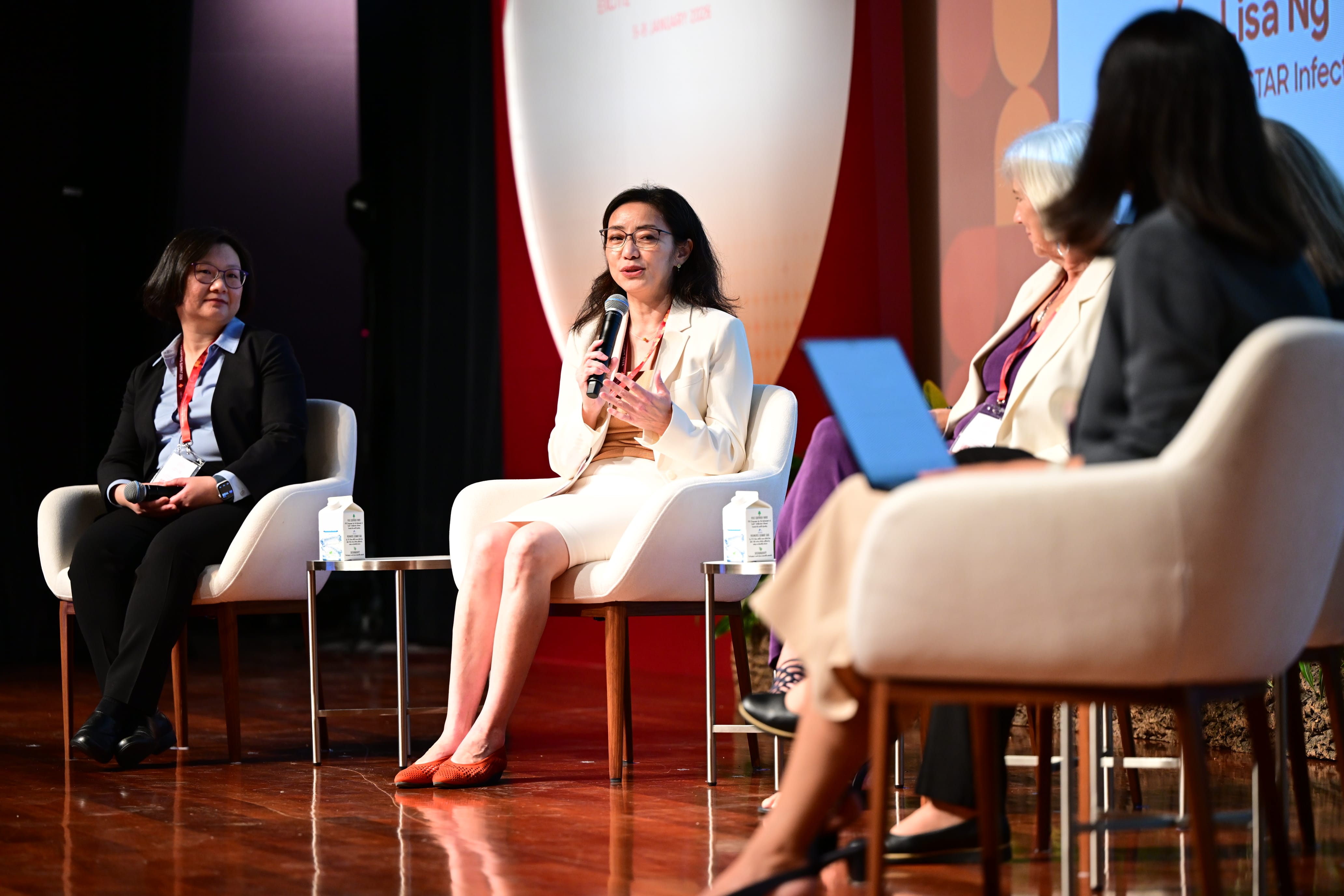Four women seated on a stage panel, with one speaking into a microphone wearing a white blazer.