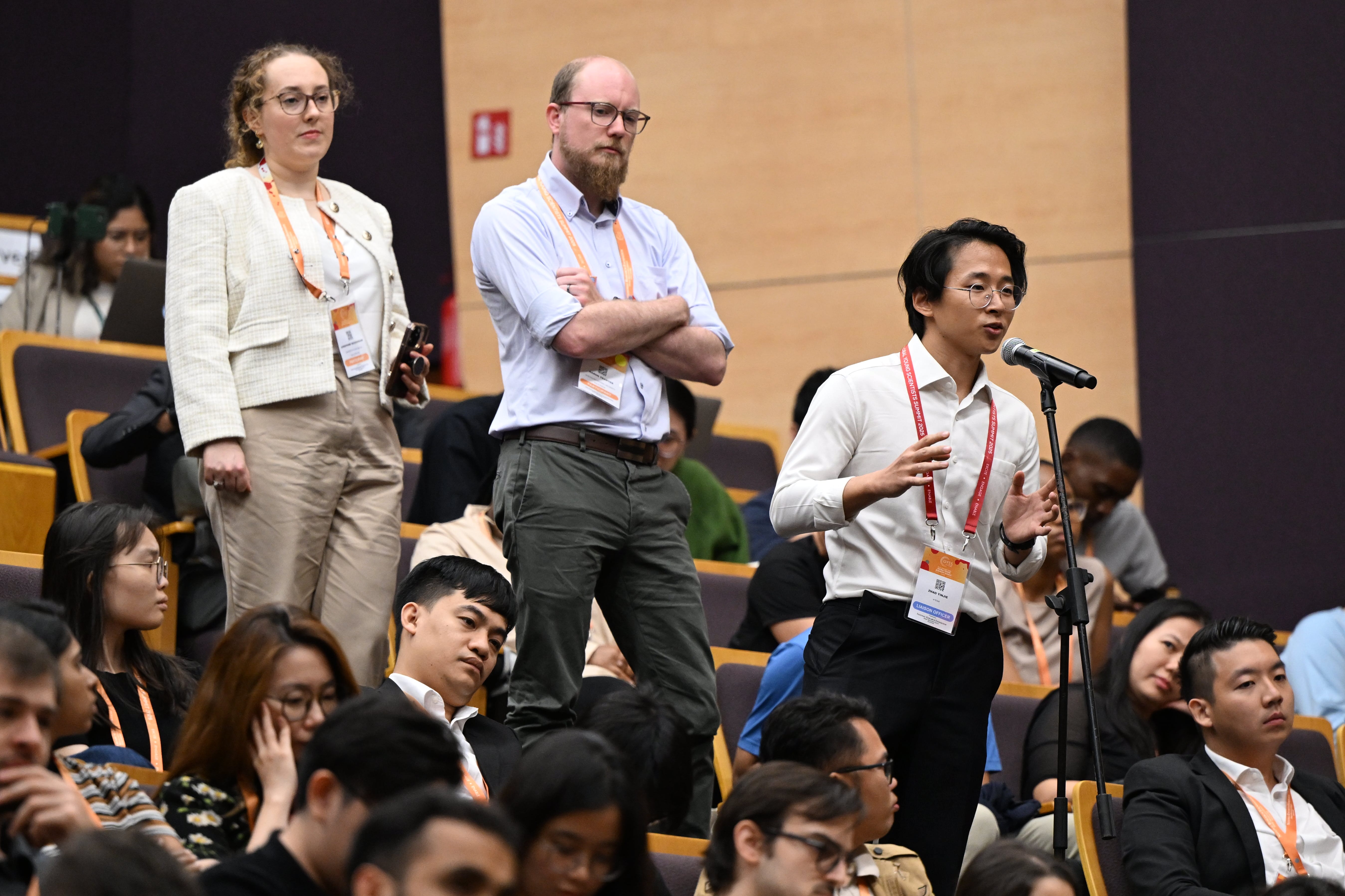 Three conference attendees stand in an auditorium, one speaking at a microphone, while others sit in rows behind them.