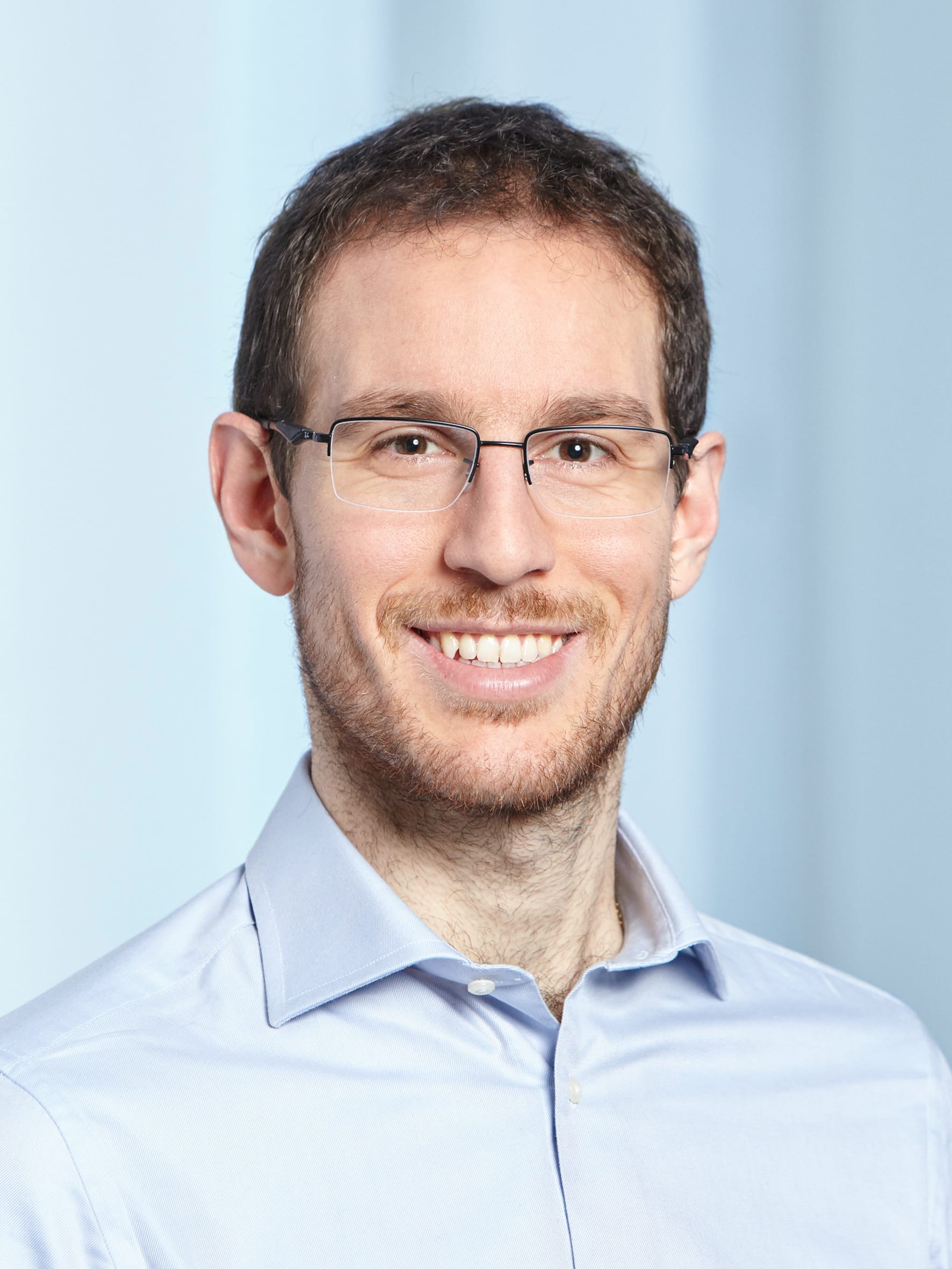 A smiling young man with dark hair, rectangular glasses, short beard, wearing a light blue shirt against a pale blue background.