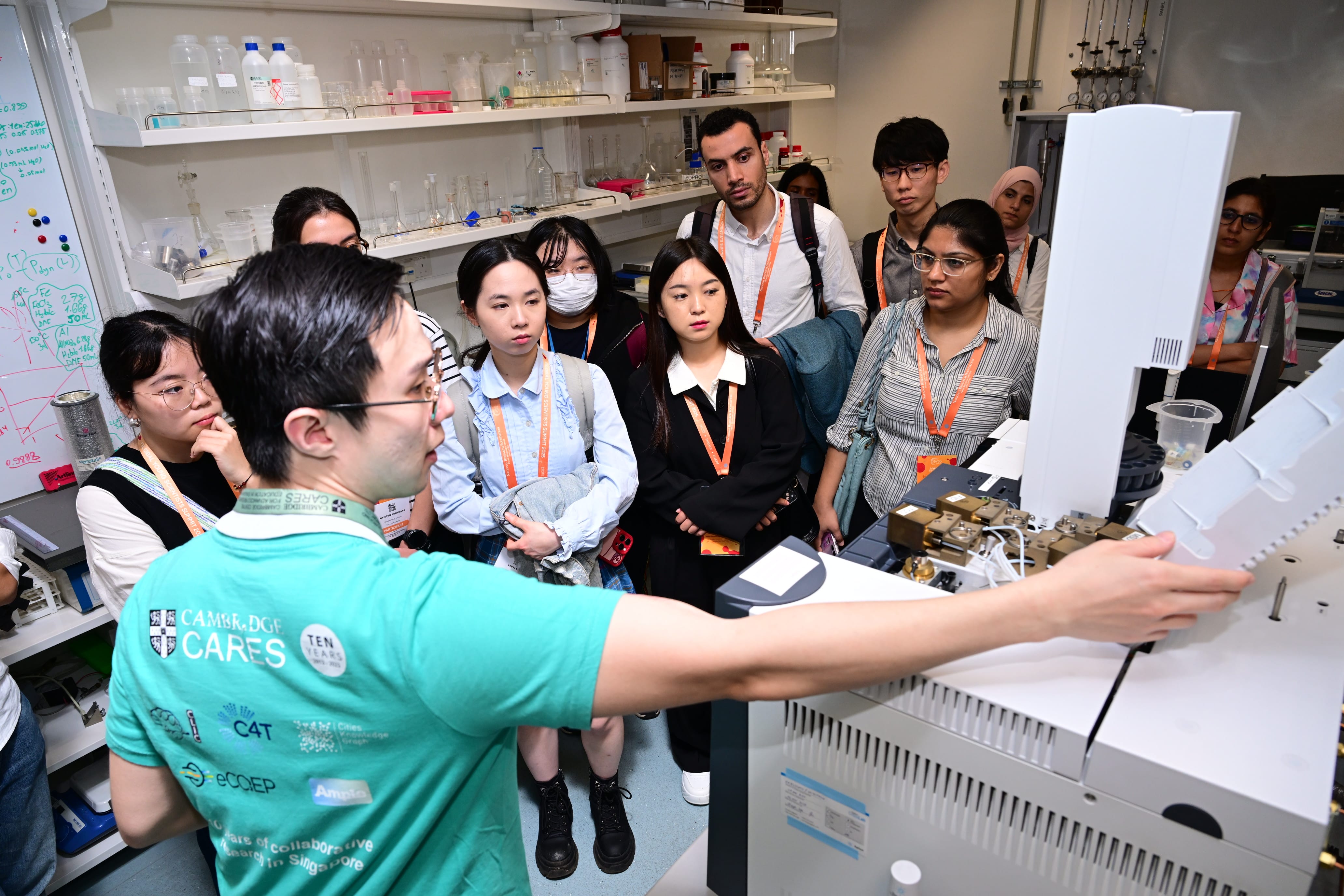A Cambridge CARES staff member demonstrates lab equipment to a group of students wearing orange lanyards in a science lab.