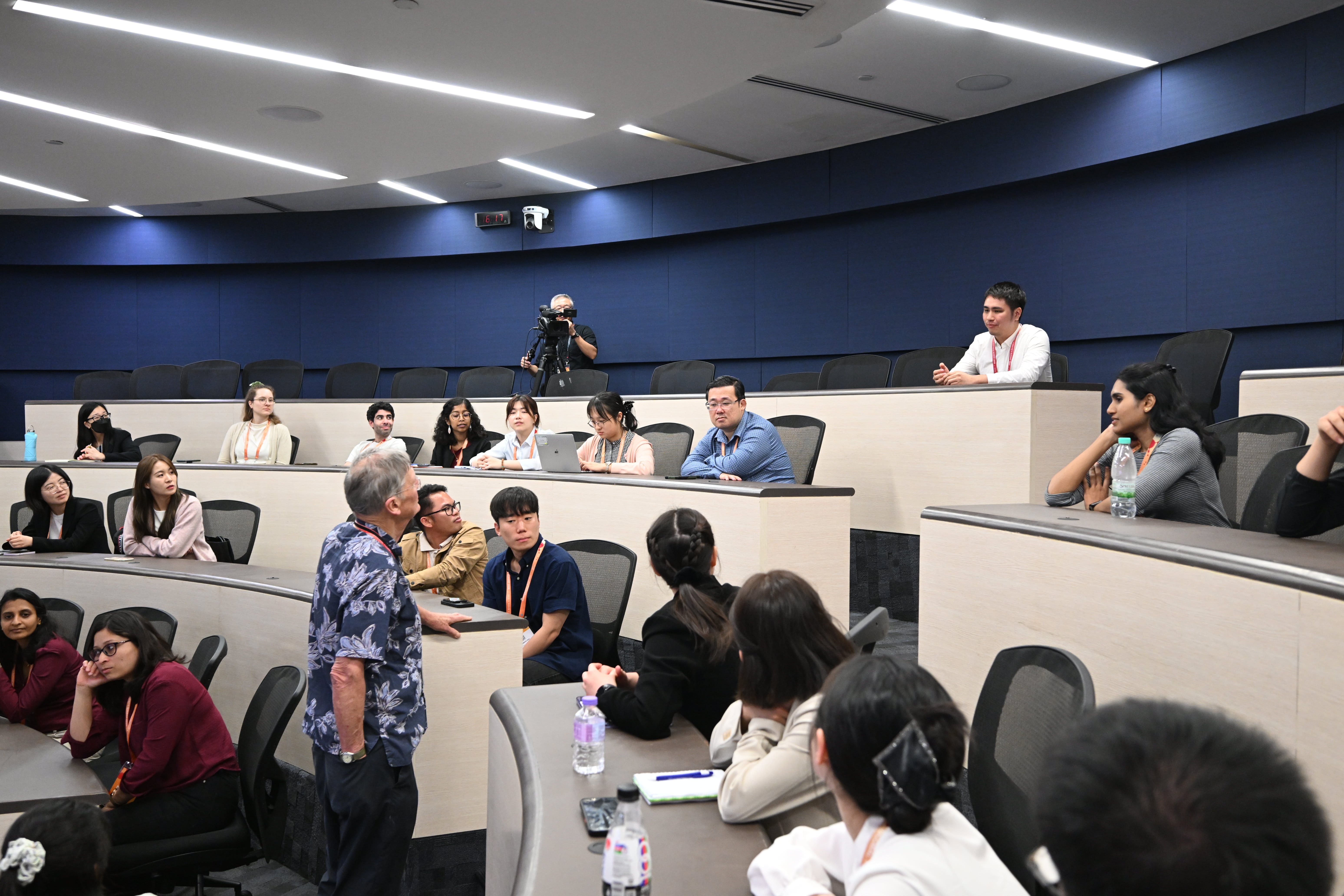 A speaker in a floral shirt talks to a group of students seated in a tiered lecture hall while a cameraman films.