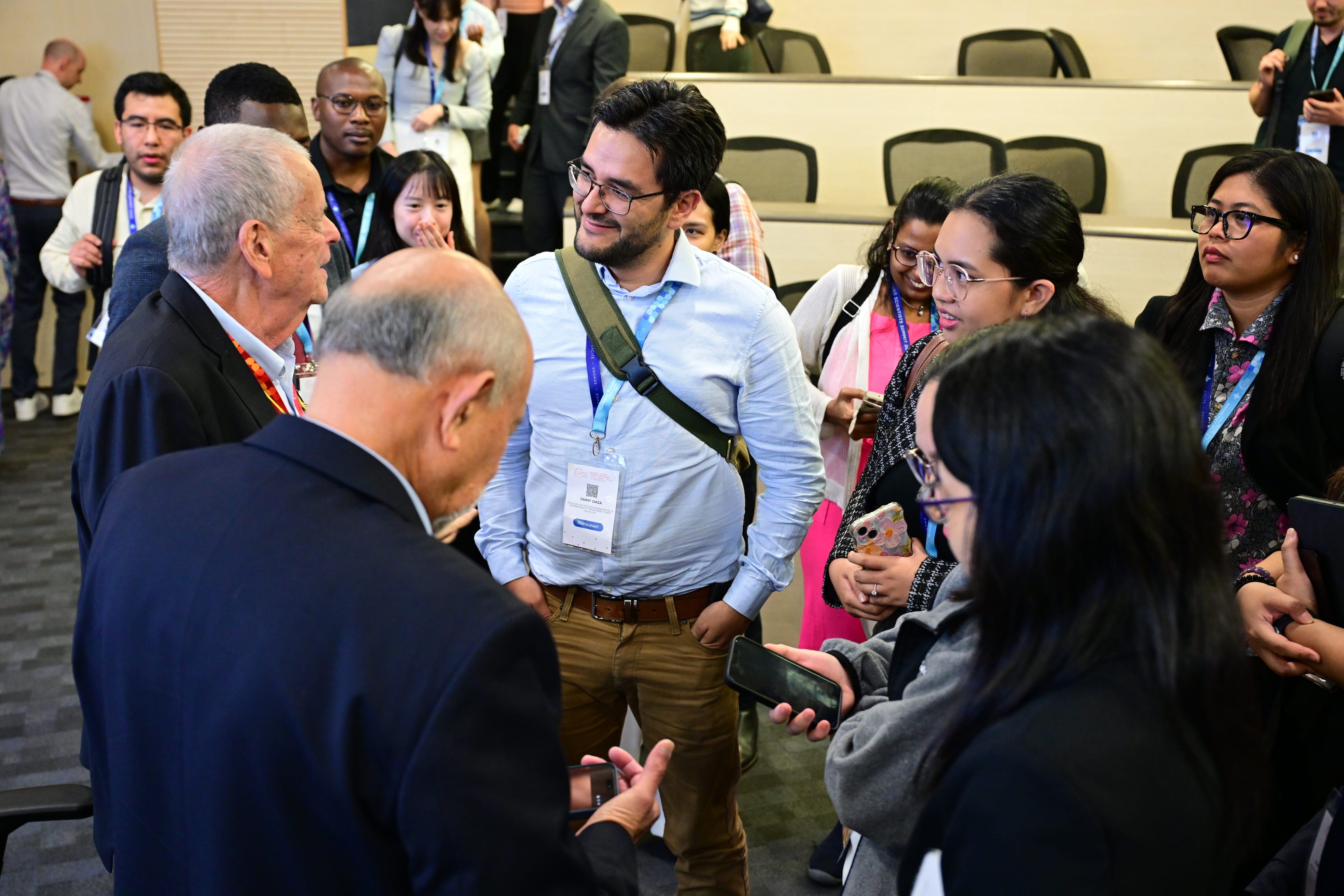 A diverse group of people wearing conference lanyards gather and converse in a meeting room with empty chairs behind them.