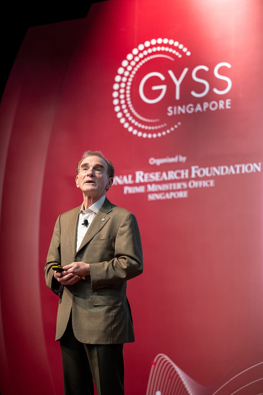 Man in a brown suit speaking on stage in front of a red GYSS Singapore banner.