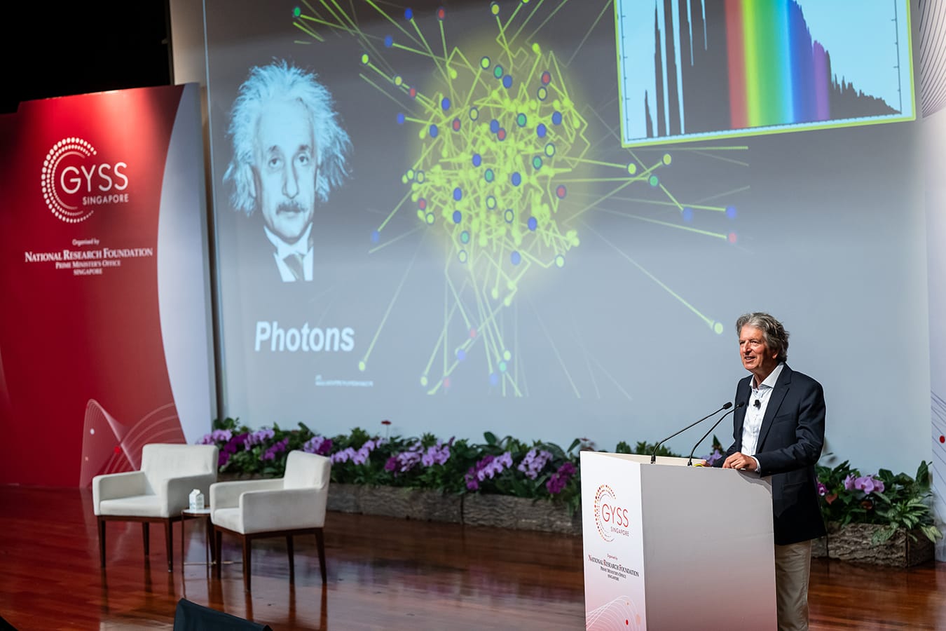 Man speaks at a GYSS Singapore podium before a slide showing Einstein's photo, a network diagram, and the word Photons.