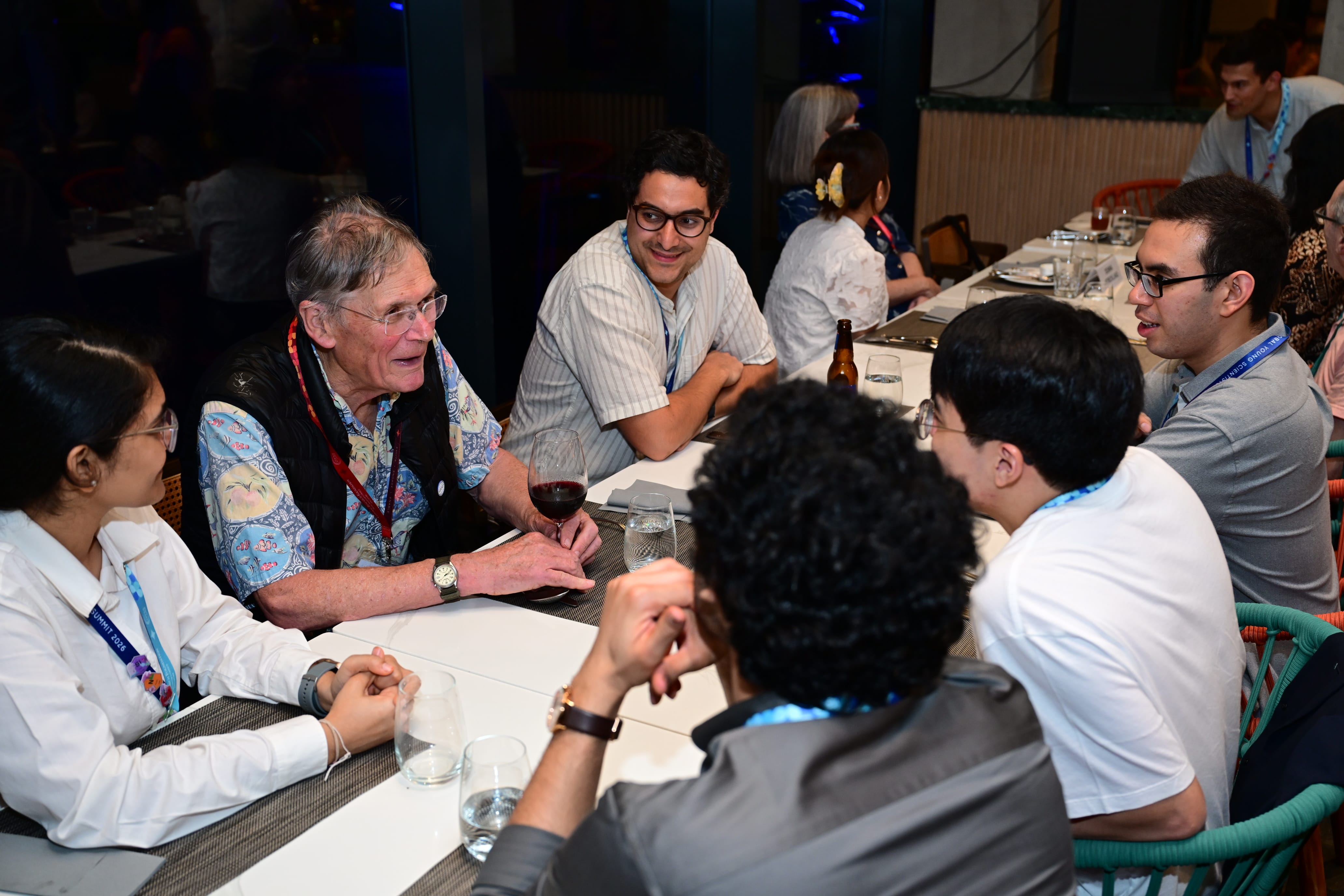 A group of people wearing lanyards sit around a table talking, with drinks visible, at an indoor event.