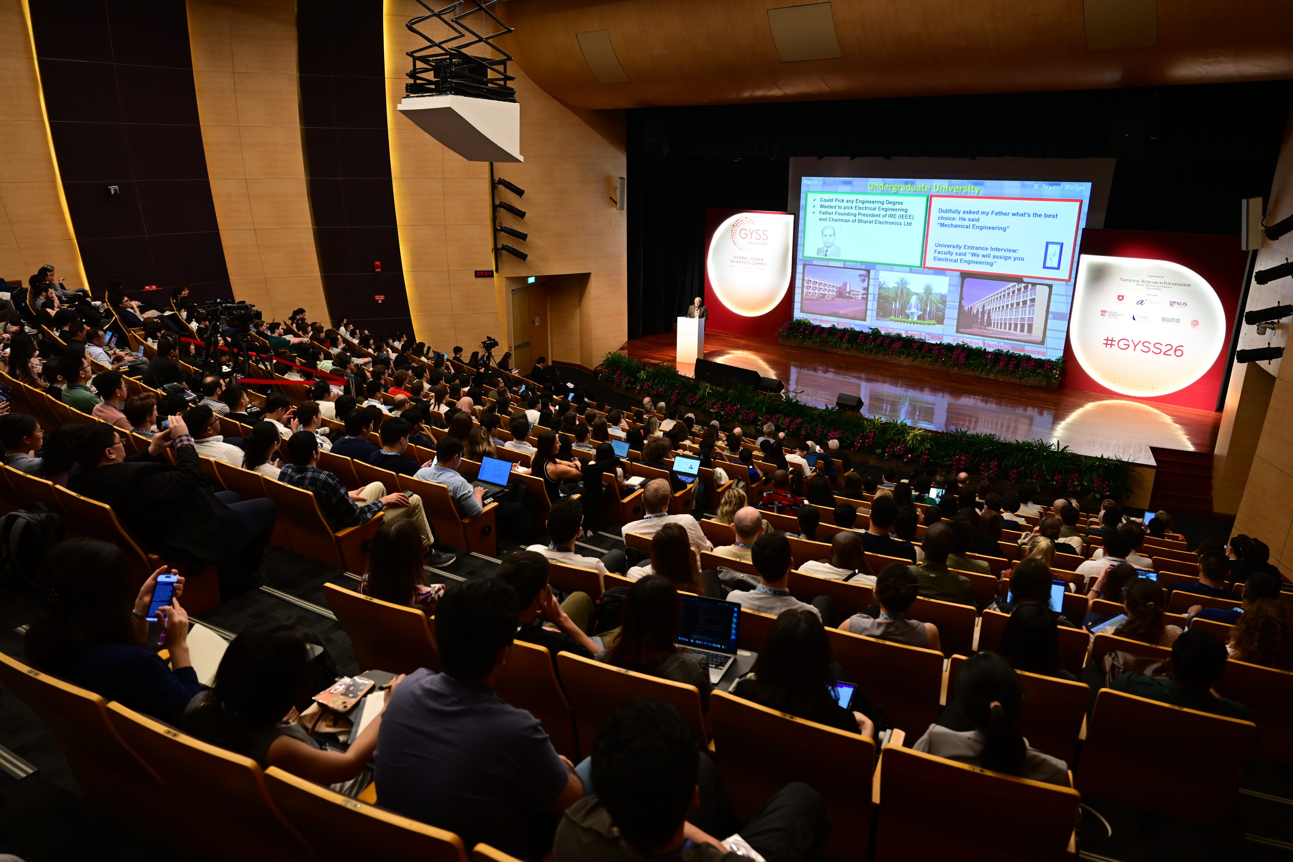 A large auditorium filled with attendees watching a speaker at a podium during the GYSS26 conference.