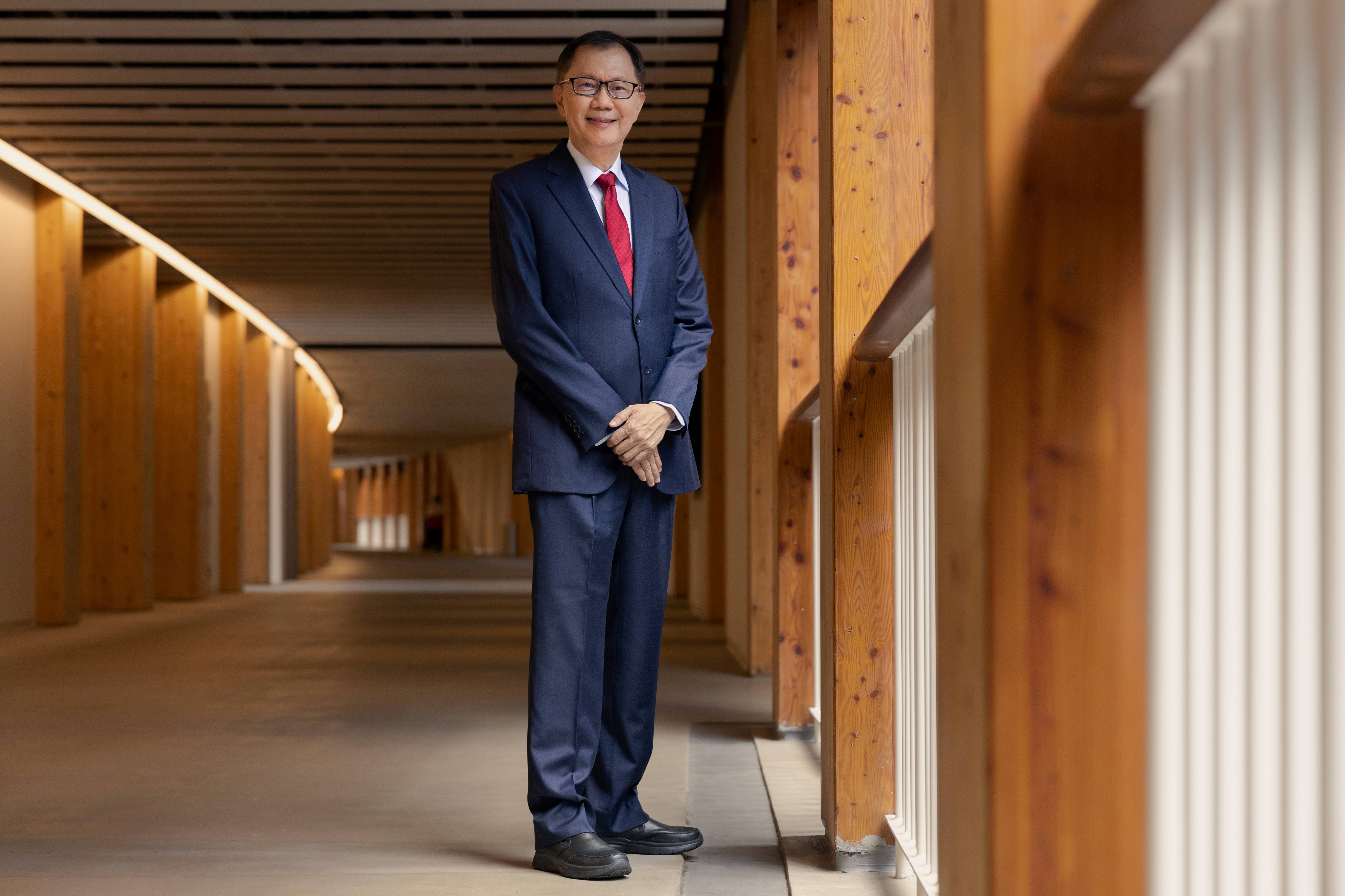 Man in a navy suit and red tie stands smiling in a long wooden-paneled corridor with warm lighting.