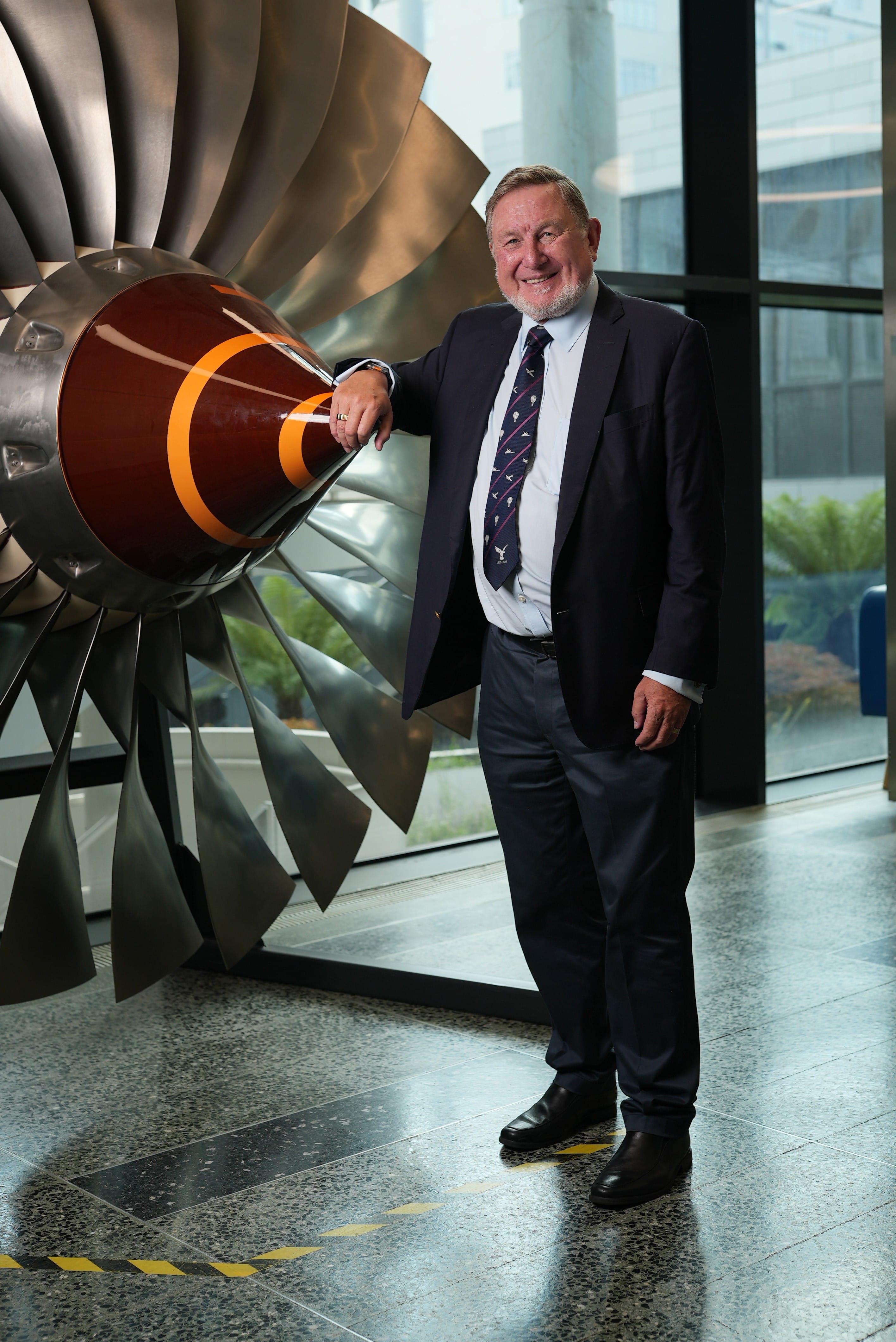 Smiling older man in a dark suit and tie leaning against a large jet engine turbine display indoors.