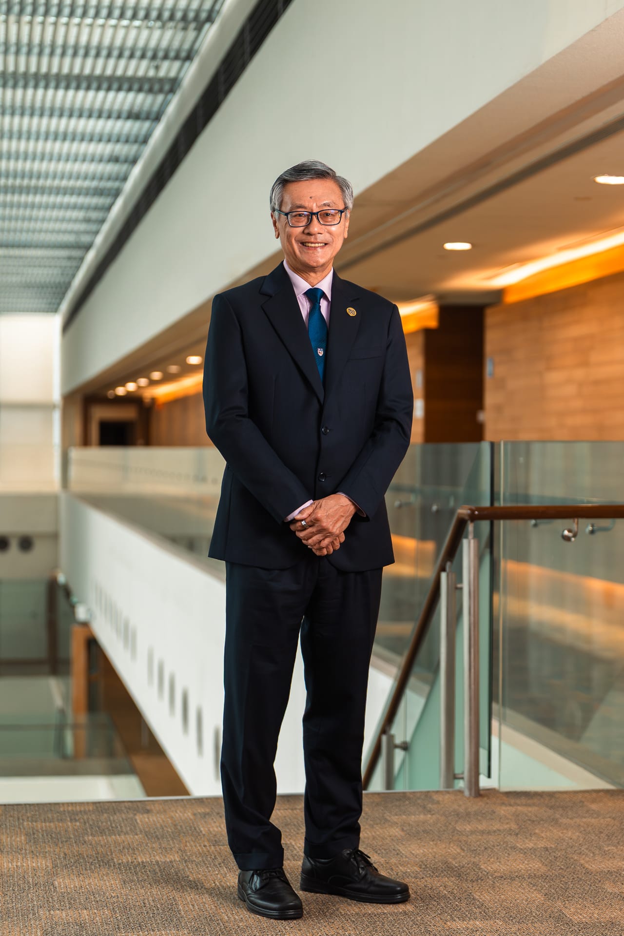 An older Asian man in a dark suit and blue tie, wearing glasses, smiling while standing in a modern building.