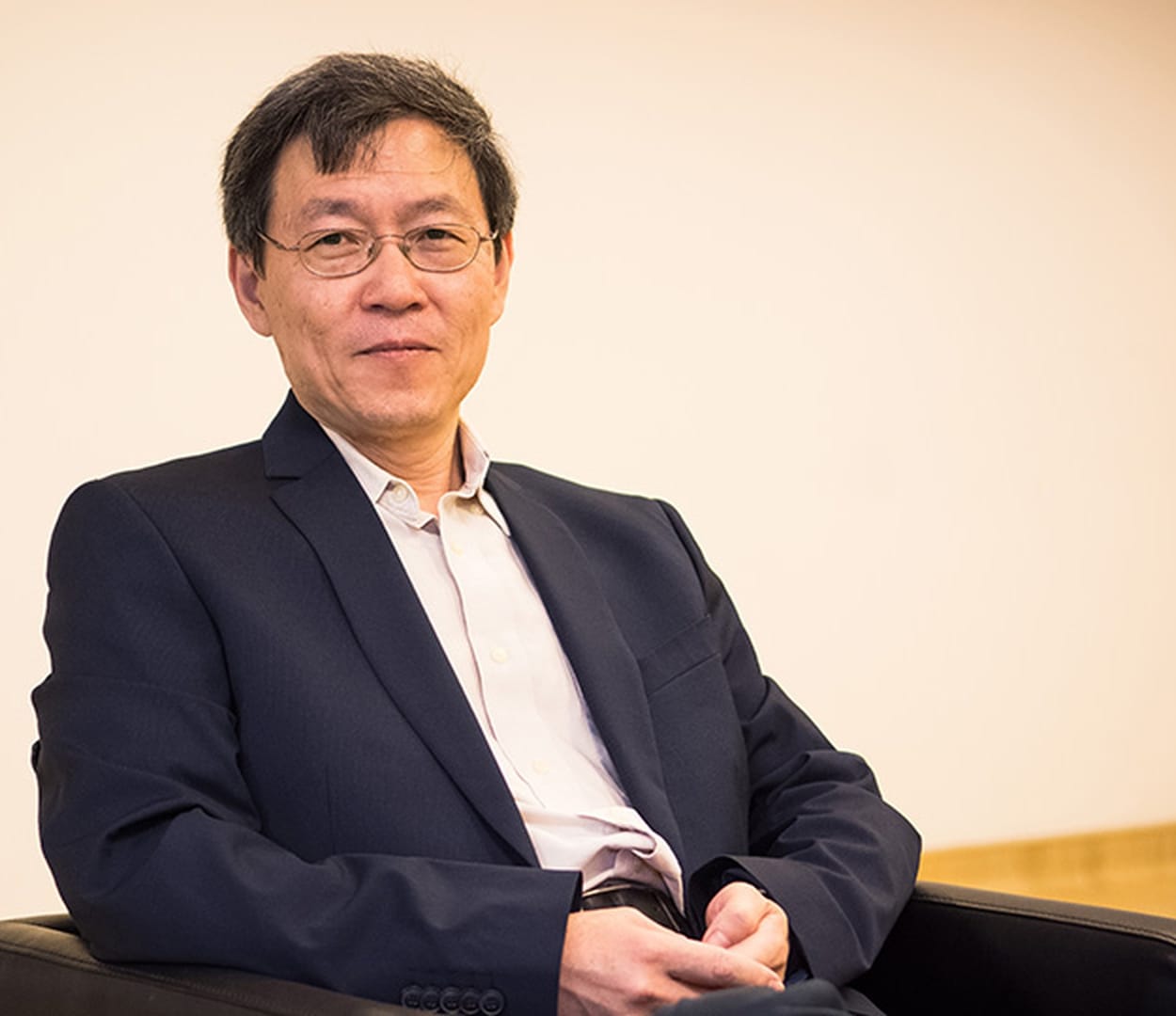 A middle-aged man with glasses wearing a dark blazer and white shirt sits with arms crossed against a plain light wall.
