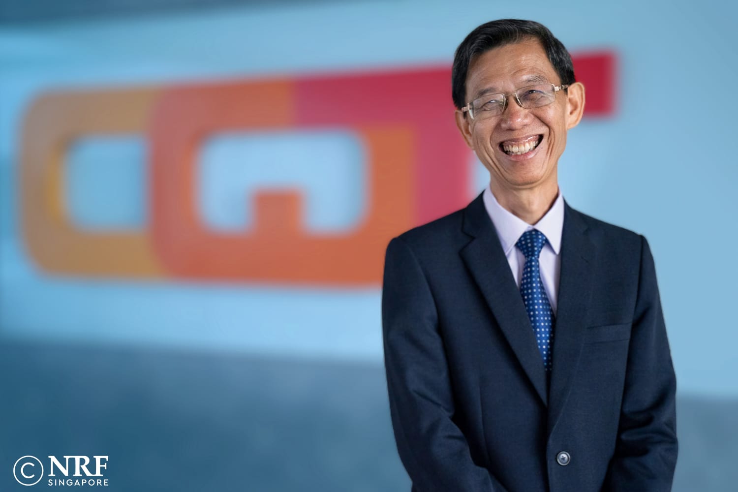 A smiling man in a dark suit and blue tie wearing glasses, standing before a blurred colorful logo background.