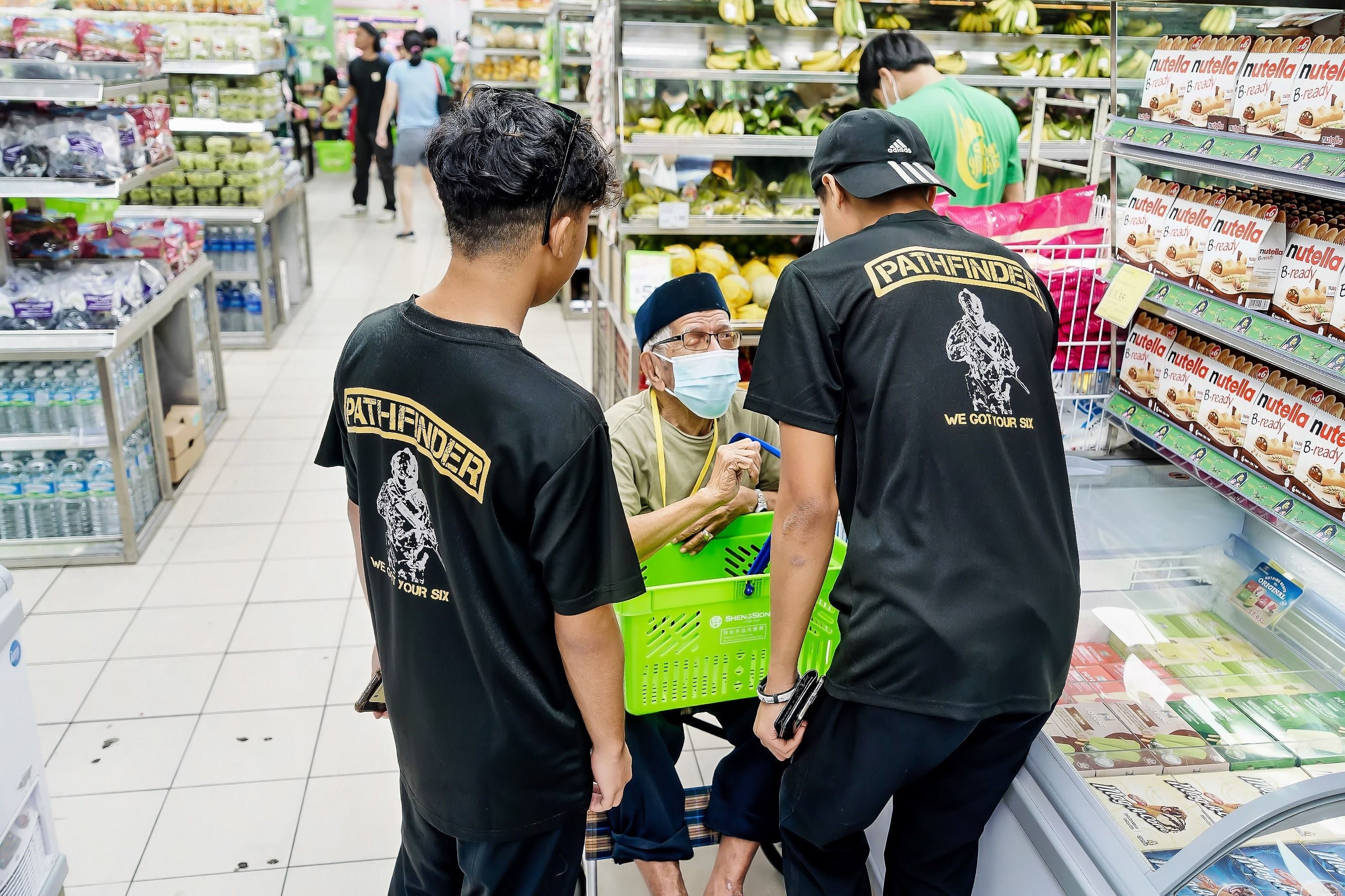 Two young men in black Pathfinder shirts assist a masked elderly man holding a green shopping basket in a grocery store.
