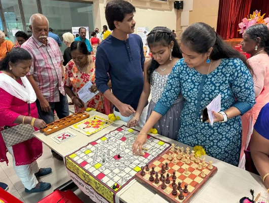 A group of people gathered around a table with a chess board and a Ludo board game at an indoor event.