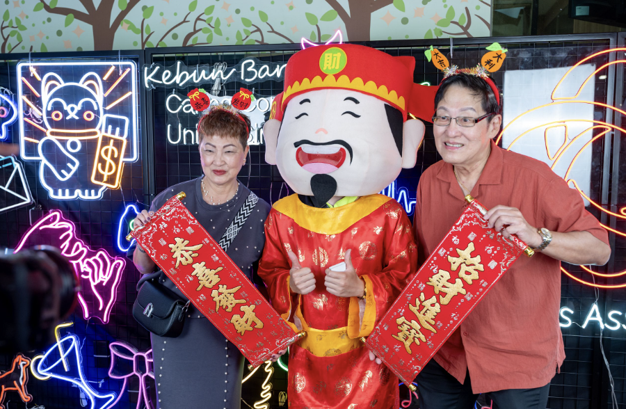 Two people hold red Chinese New Year banners beside a large God of Fortune mascot, with neon decorations behind them.