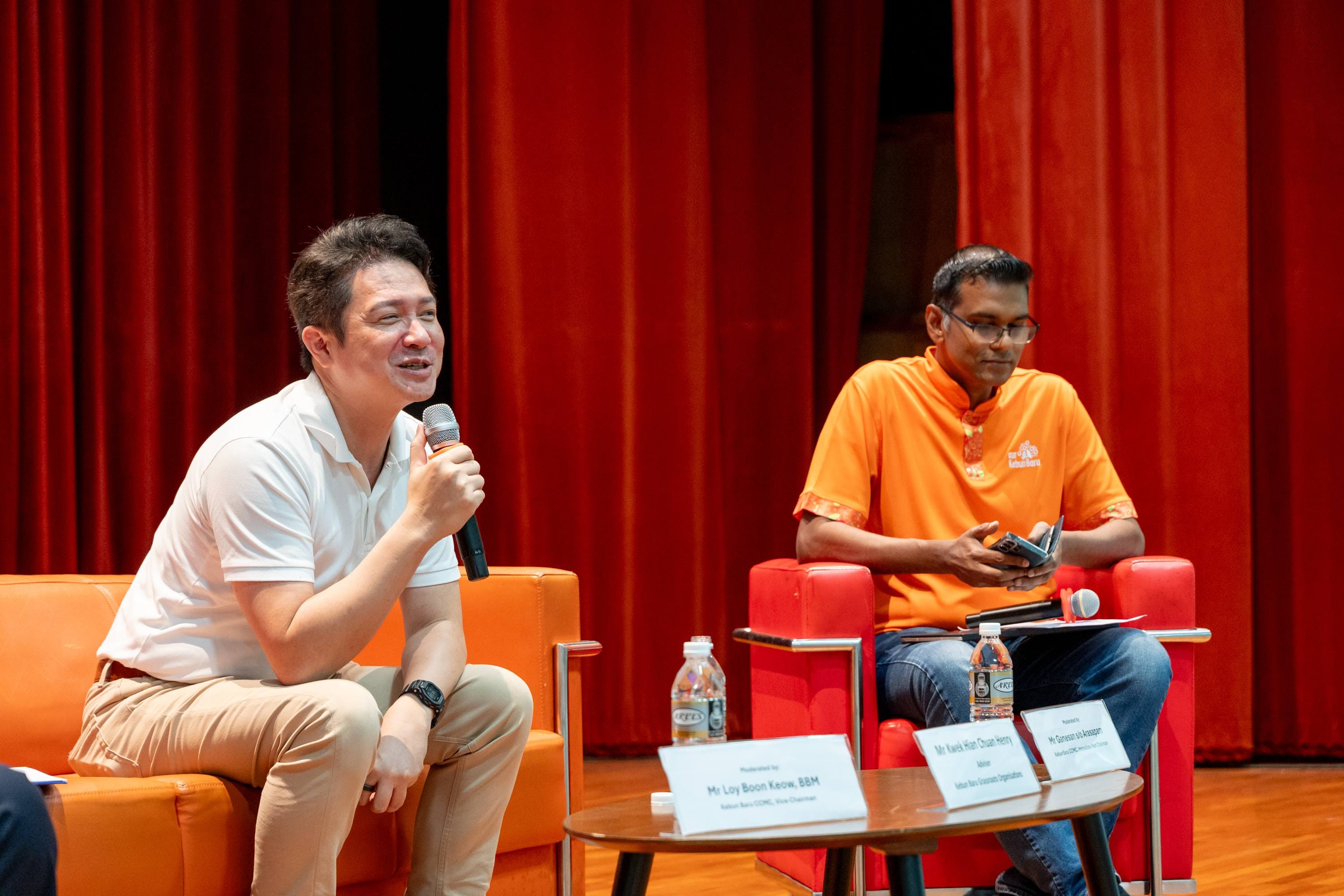 Two men sit on stage during a panel; one speaks into a microphone, the other in an orange shirt listens.