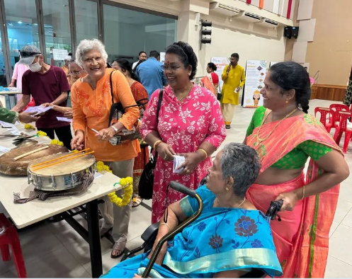 Four women in colorful traditional Indian attire gather around a drum; one woman sits in a wheelchair.