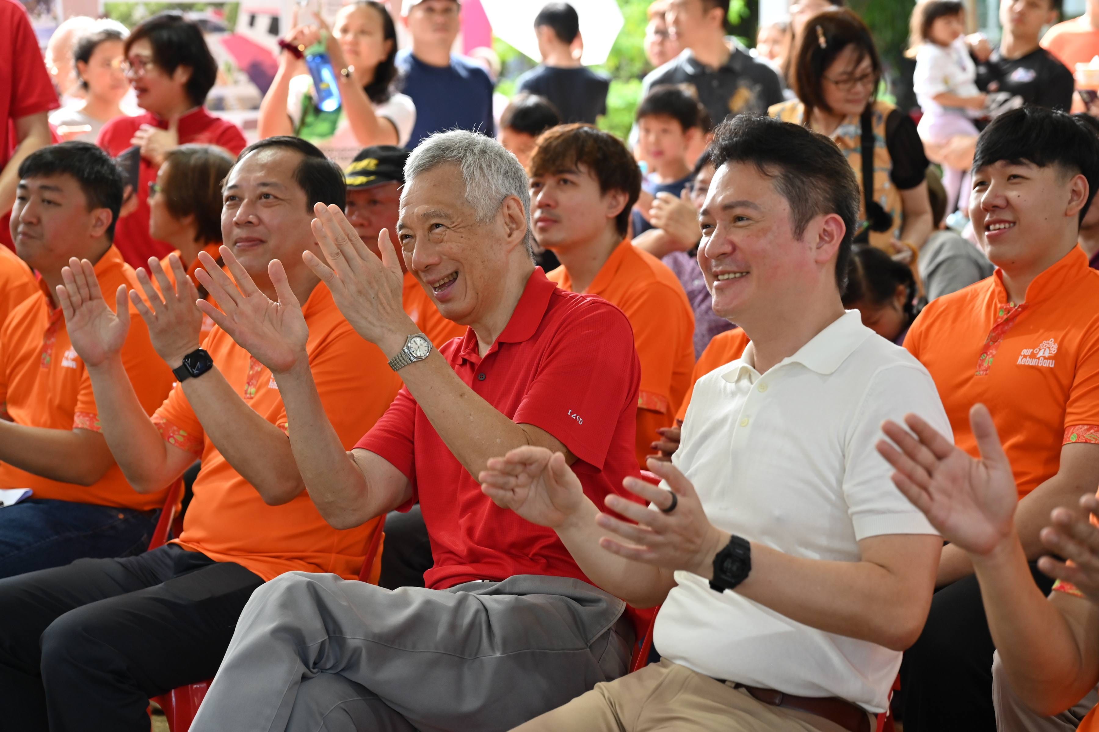 A smiling older man in red and others in orange shirts clap and cheer at an outdoor community event.