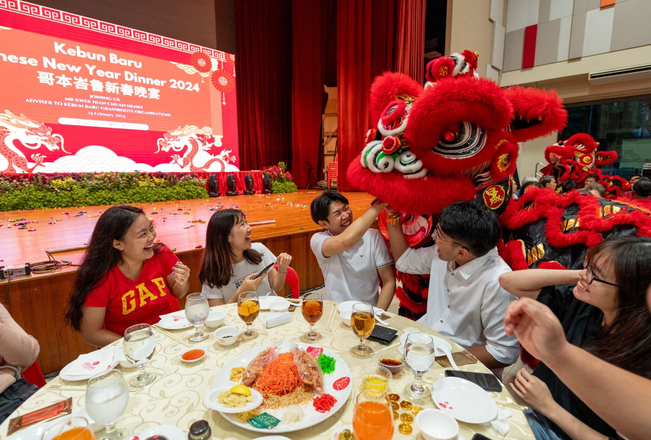 Young people laugh at a dinner table as a red lion dance costume approaches at a 2024 Chinese New Year event.