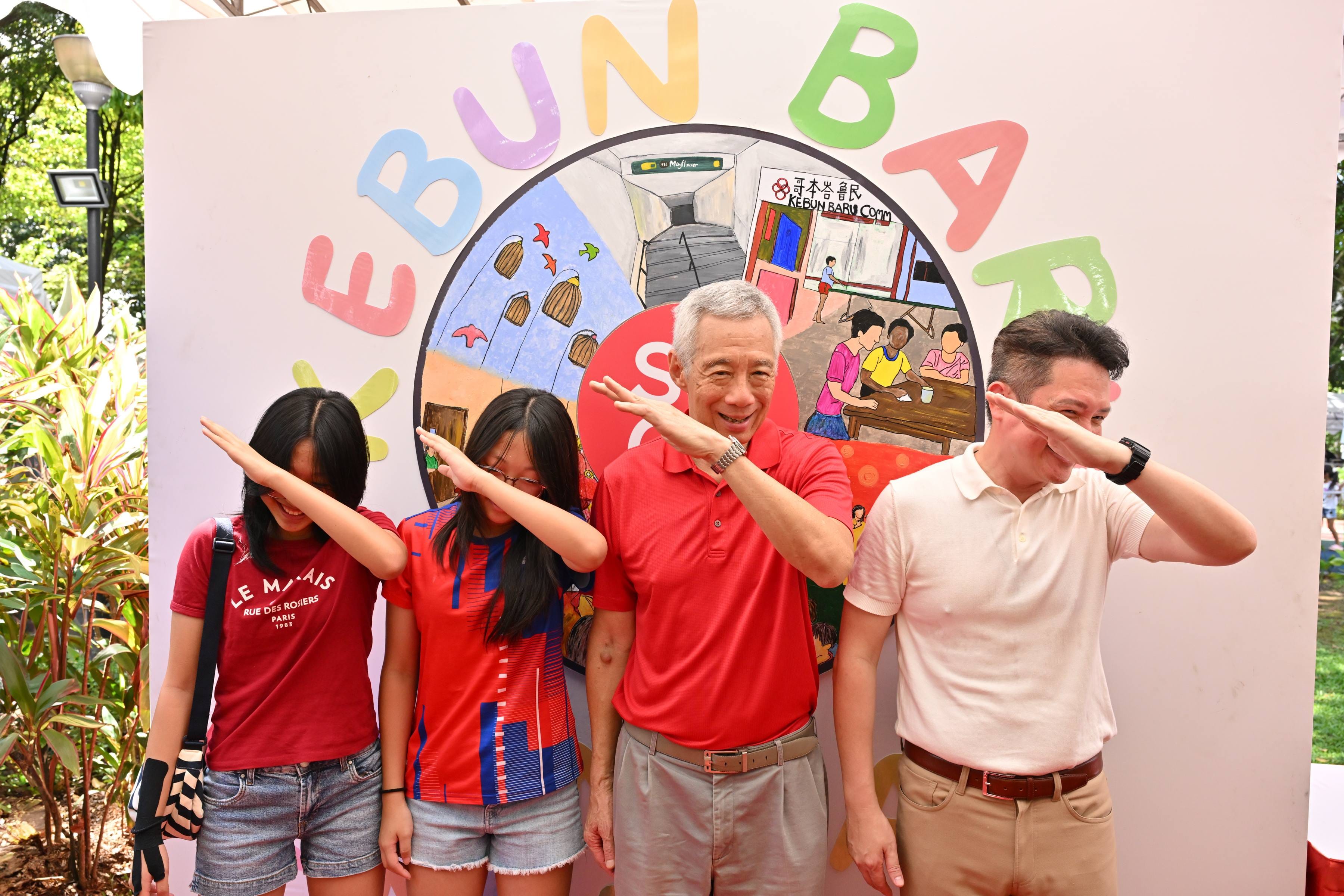 Four people pose in front of a colorful Kebun Baru mural, two girls dabbing, an older man and a younger man saluting.