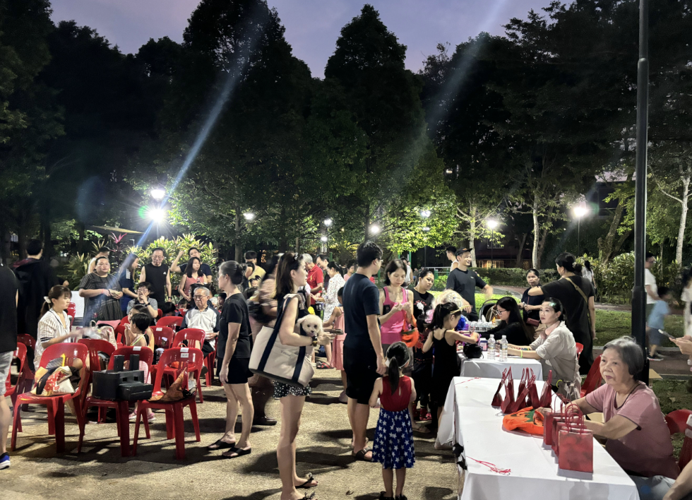 A crowd of people gathered outdoors at night around tables with red chairs, lit by park lights and surrounded by trees.