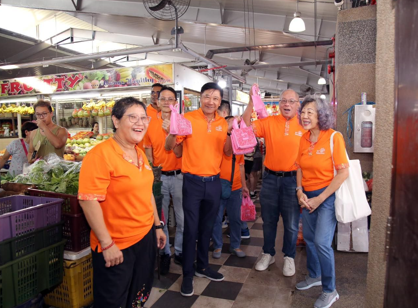 A group of smiling adults in matching orange polo shirts hold up pink bags inside a busy indoor market.