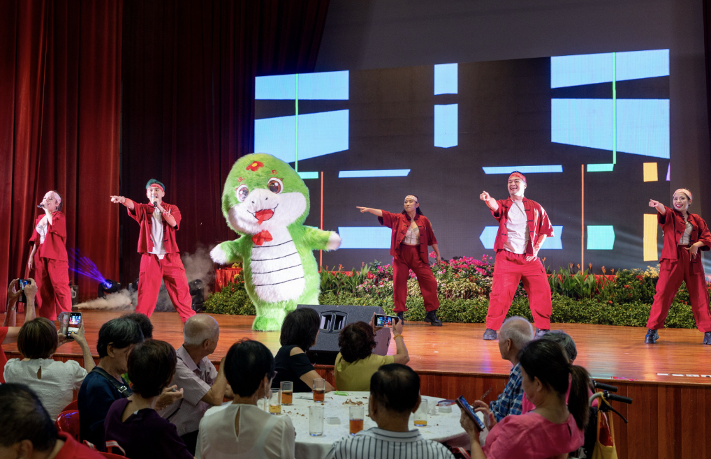 Performers in red outfits dance on stage with a green cartoon snake mascot costume while an audience watches and records.