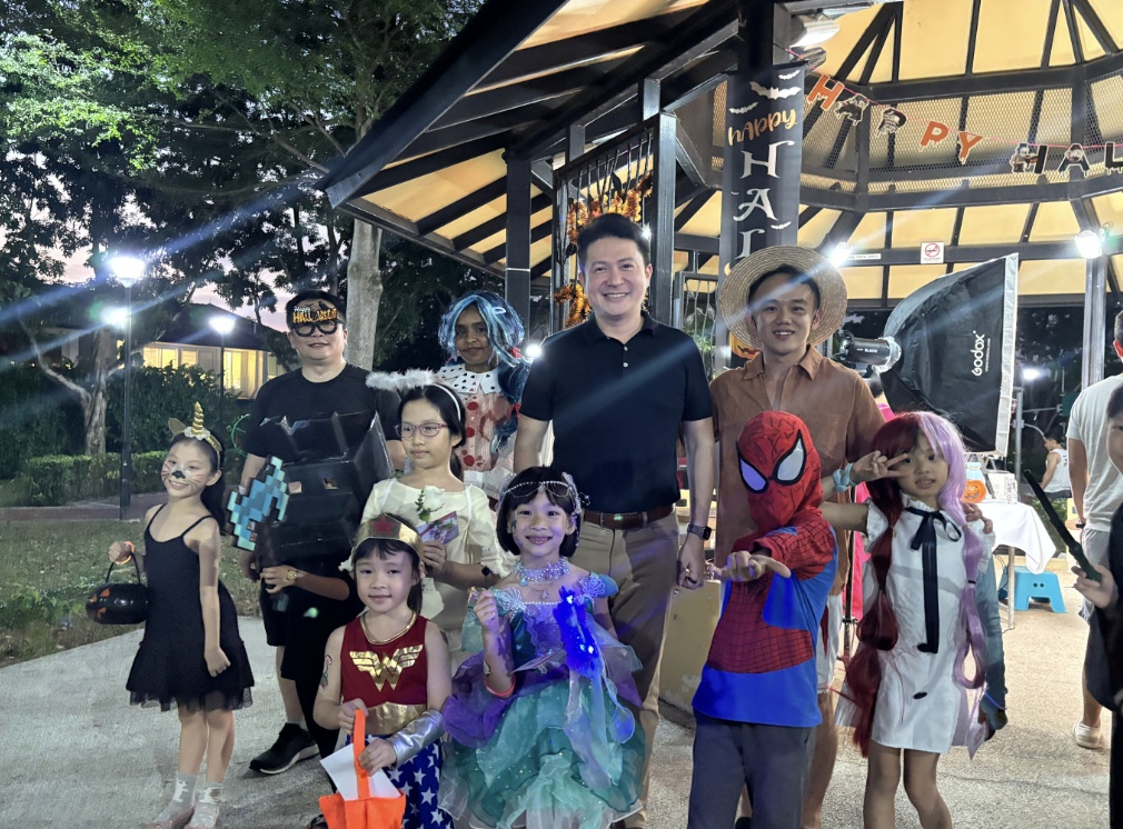 Group of adults and children in Halloween costumes posing outdoors under a Happy Halloween banner at night.