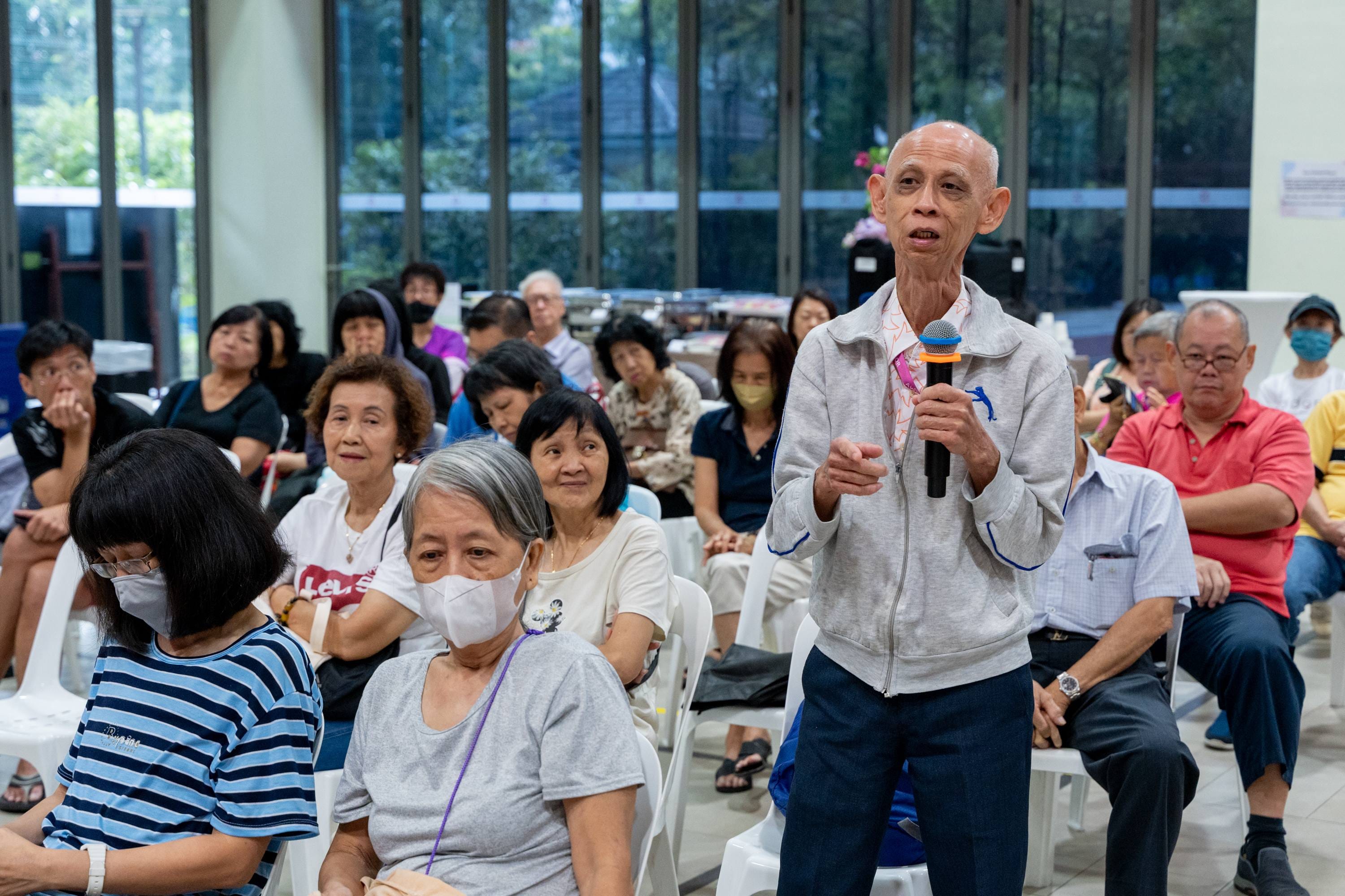 An elderly man in a grey jacket stands holding a microphone, speaking to a seated crowd of older adults indoors.