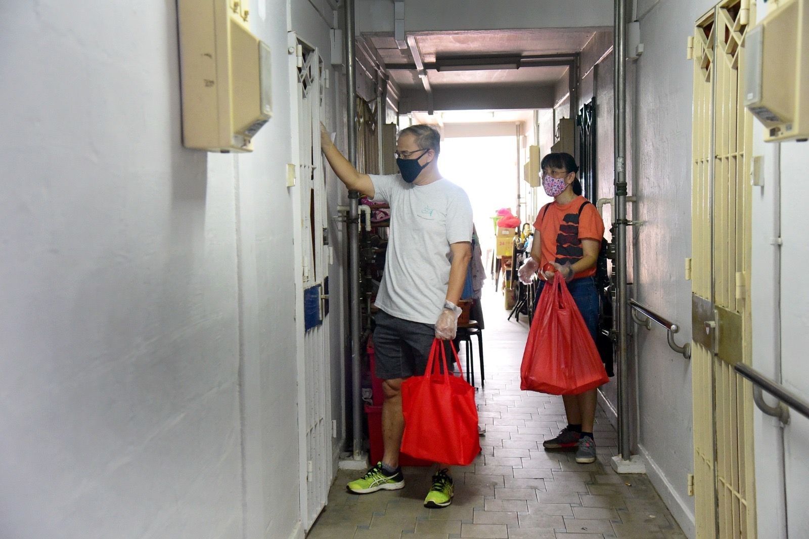 Two masked people carrying red bags knock on a door in a narrow apartment corridor.