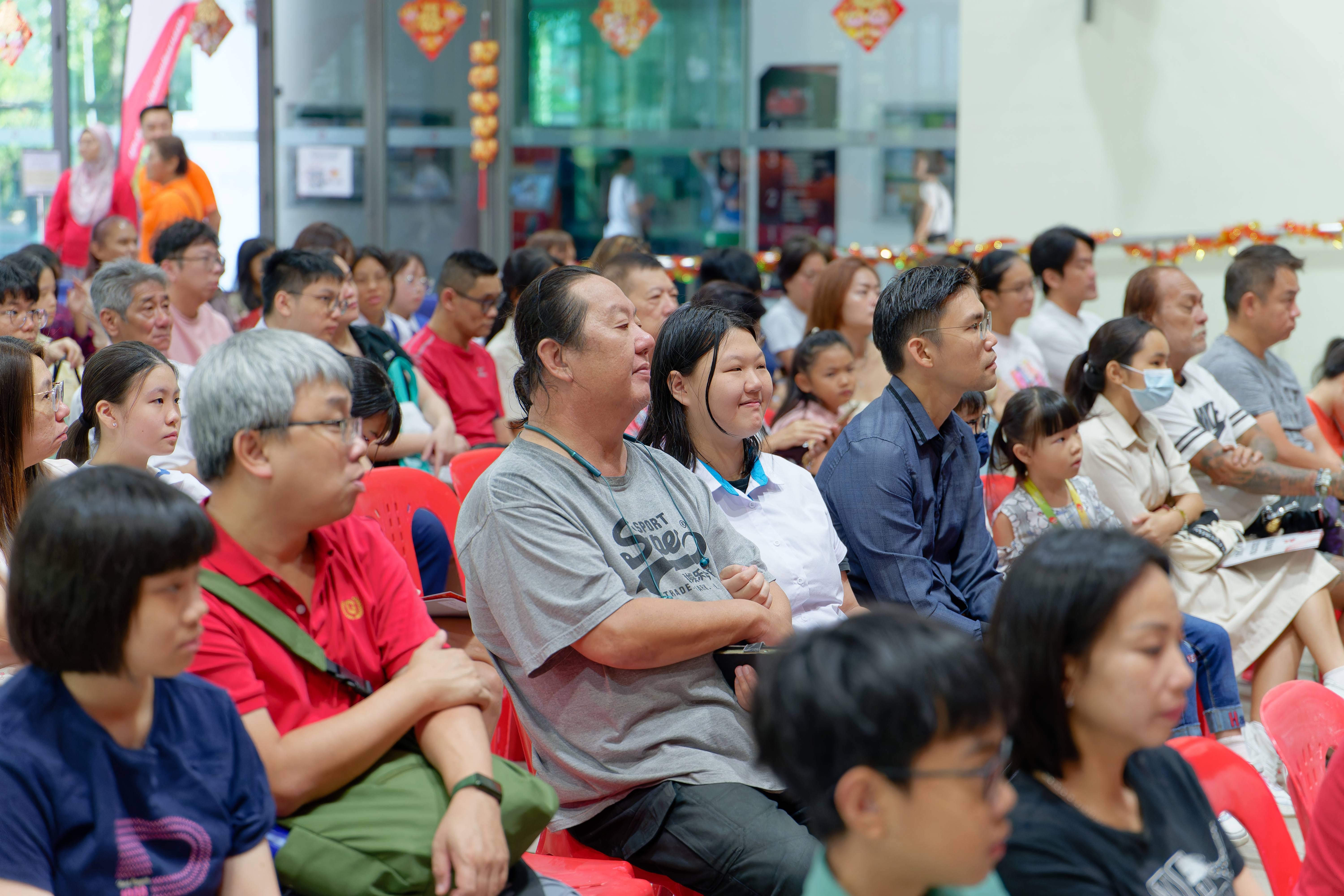 A large crowd of adults and children seated on red chairs at an indoor event with Chinese New Year decorations.