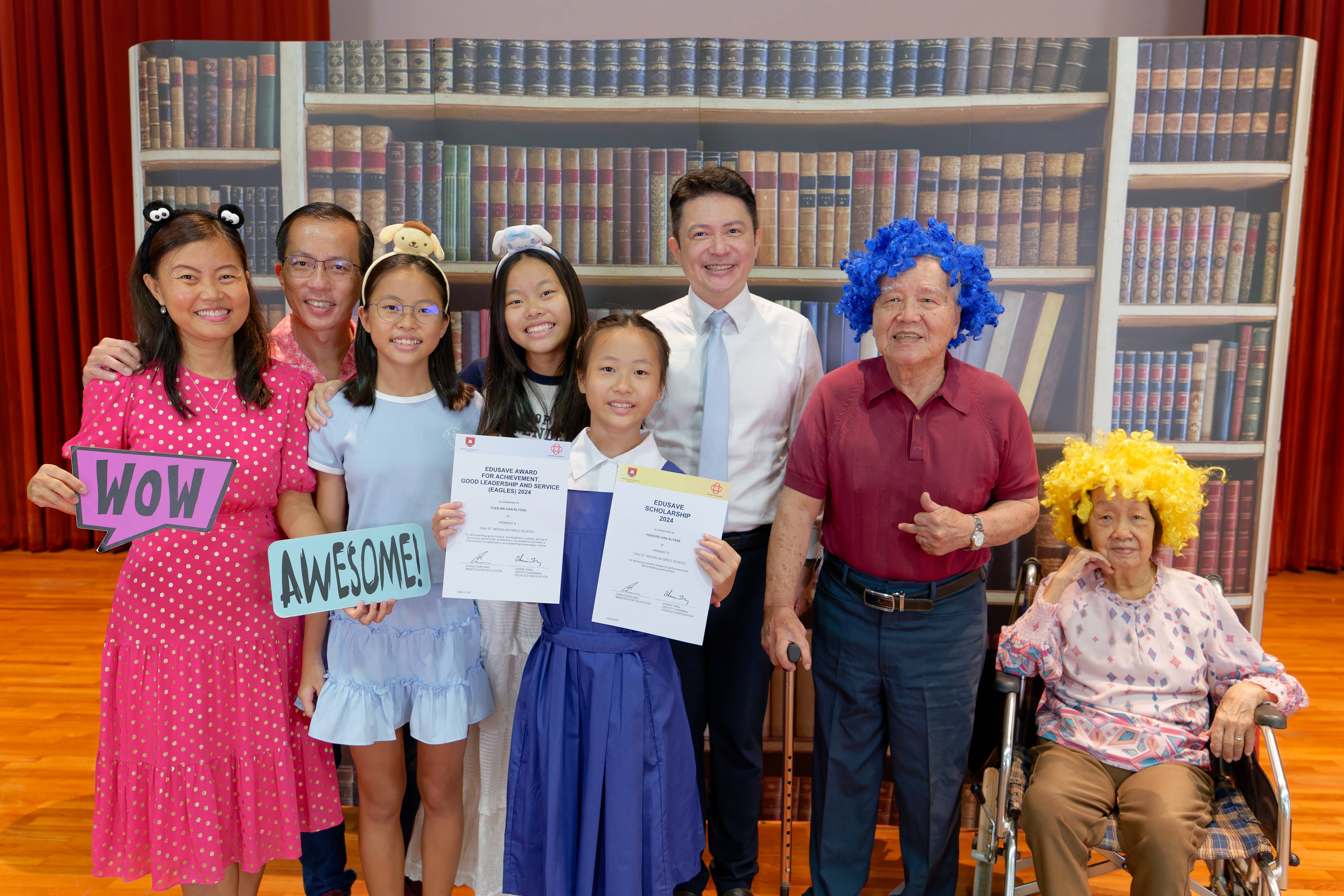 Family group posing with Edusave award certificates, fun wigs, and WOW and AWESOME signs in front of a bookshelf backdrop.