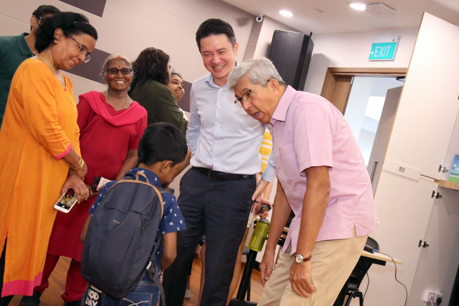 A group of adults and a child with a backpack gather indoors near an exit sign, smiling and interacting.