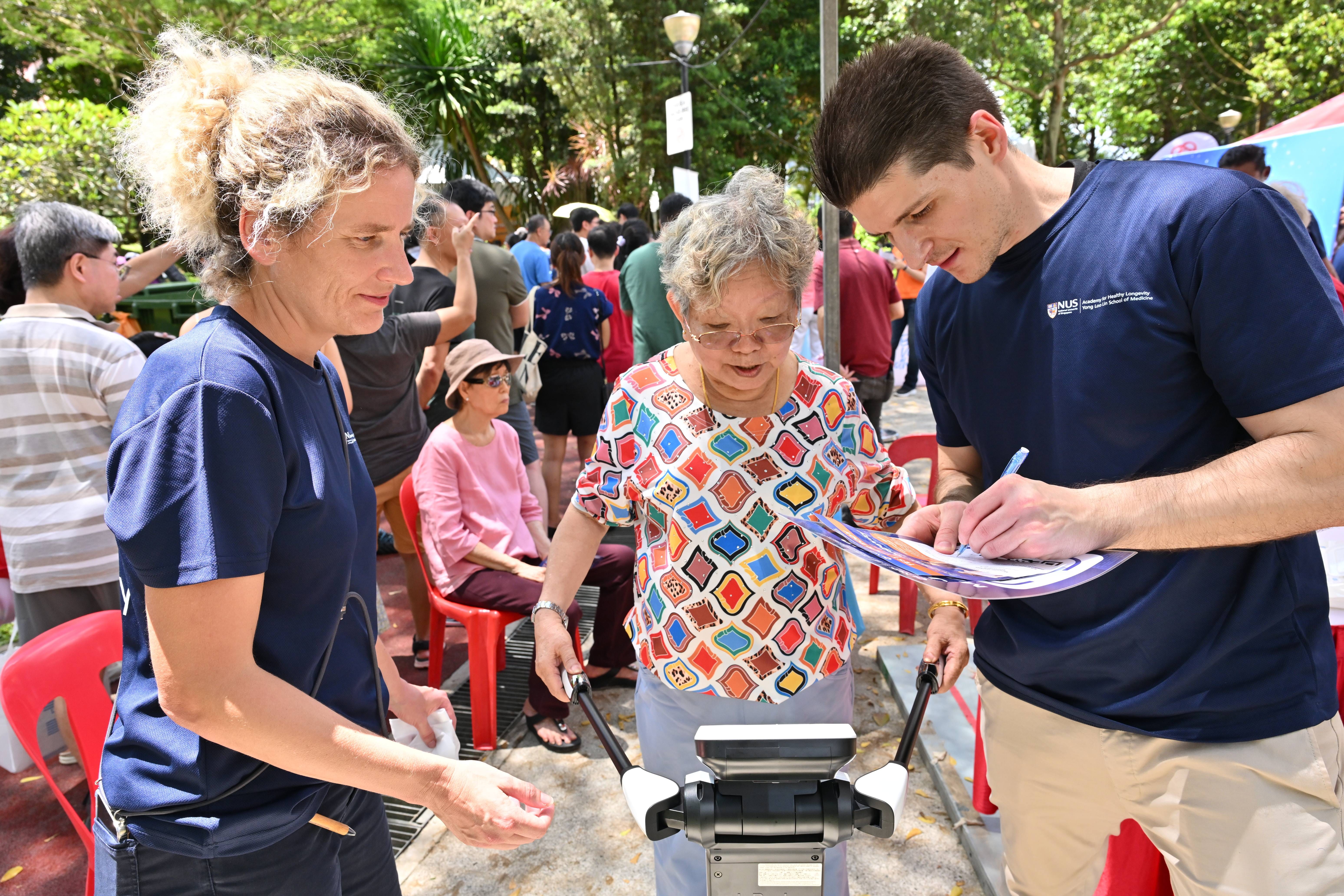 Two NUS staff in navy shirts assist an elderly woman with a device at an outdoor community health event.