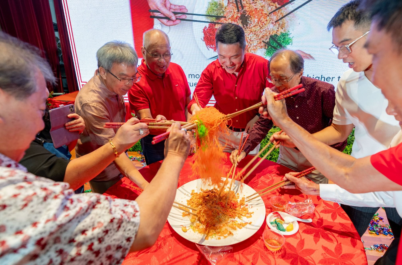 Six people use chopsticks to toss colorful yusheng salad high above a white plate on a red tablecloth.