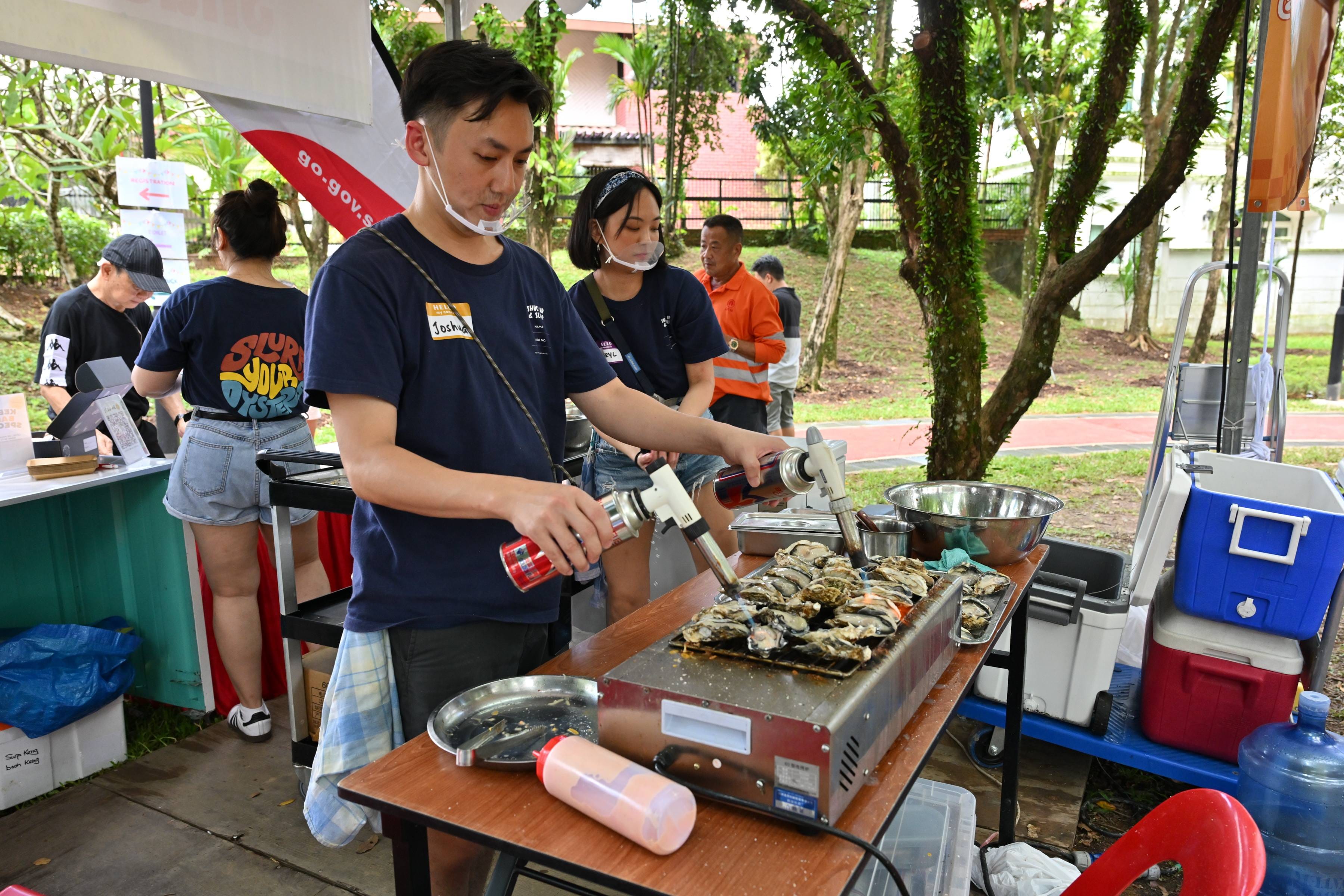 Two people in navy shirts use blowtorches to prepare oysters on a grill at an outdoor food stall.